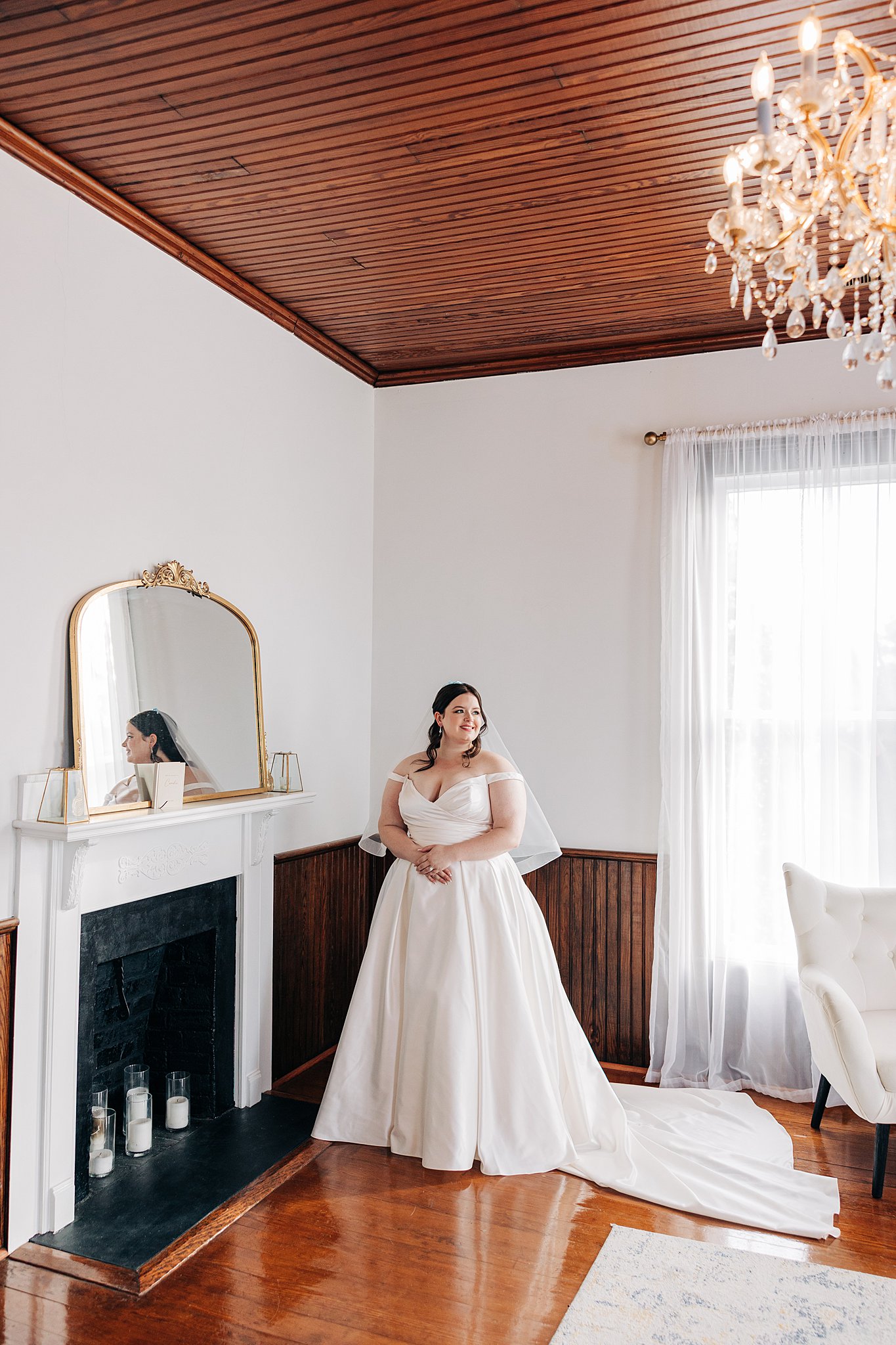 A bride stands in a getting ready room by a fireplace smiling over her shoulder