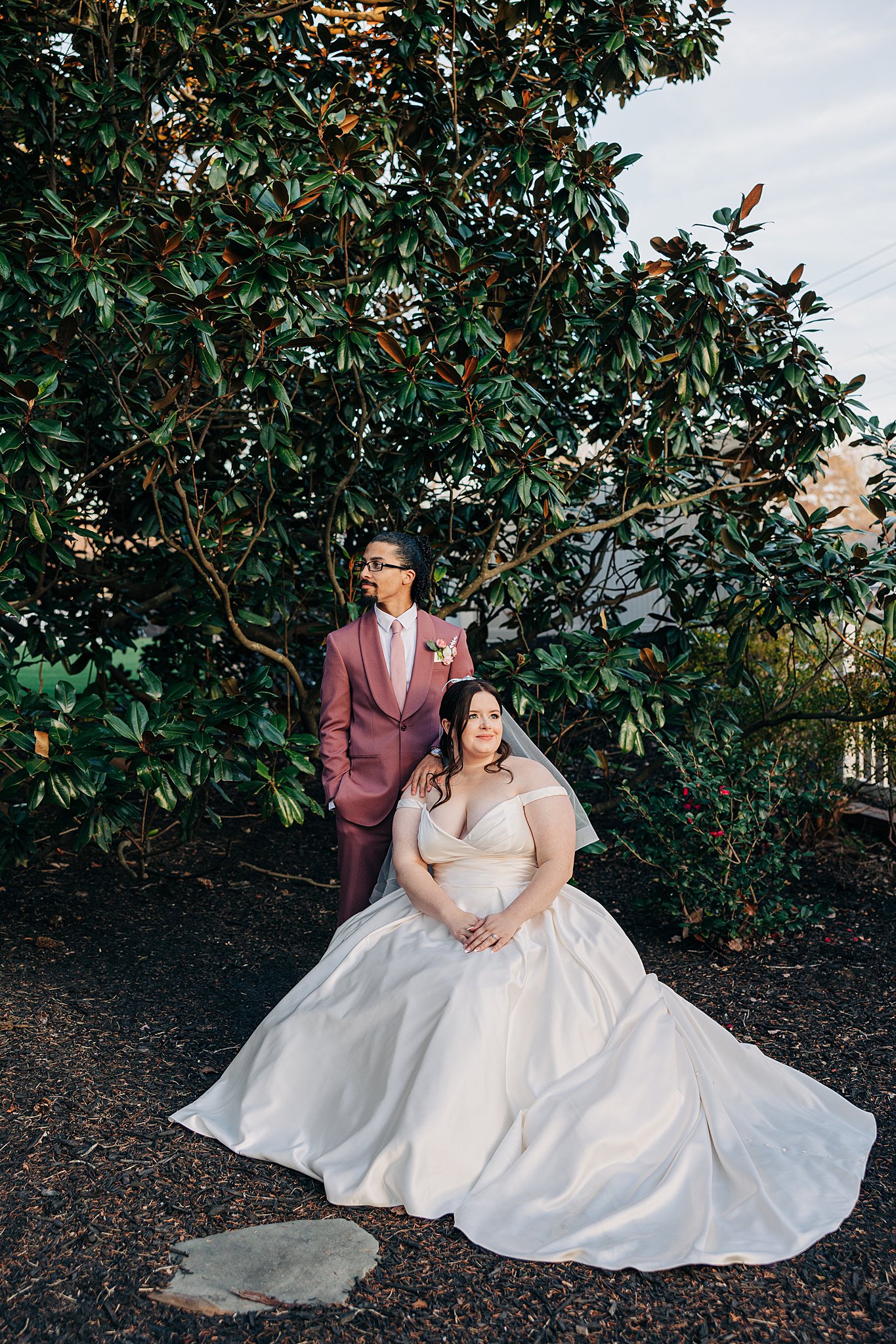A bride in a silk dress sits on a garden bunch with her groom in a pink suit standing with a hand on her shoulder at The Victorian: Youngsville