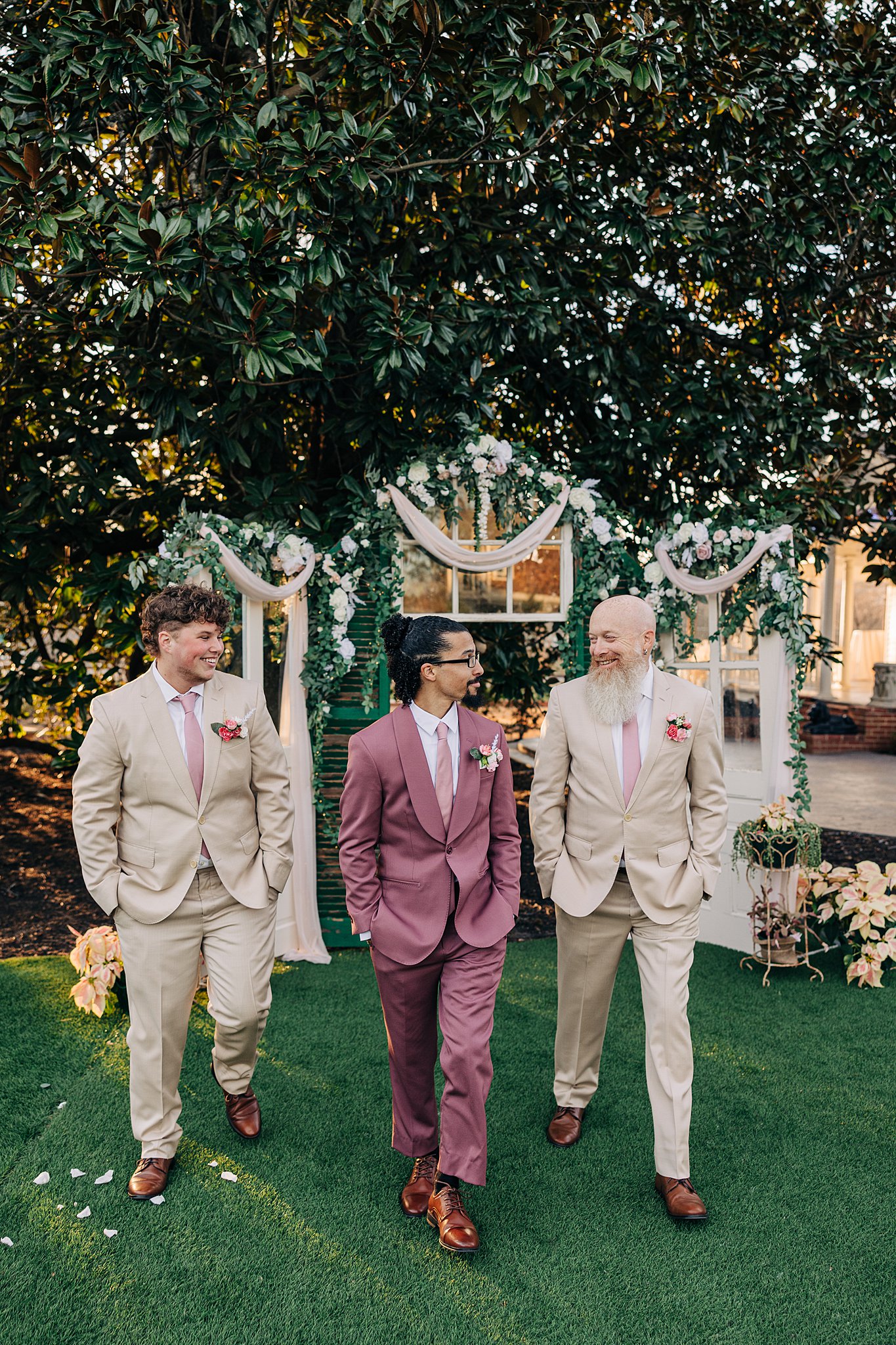 A groom in a pin suit walks in a garden with hands in his pockets with his groomsmen