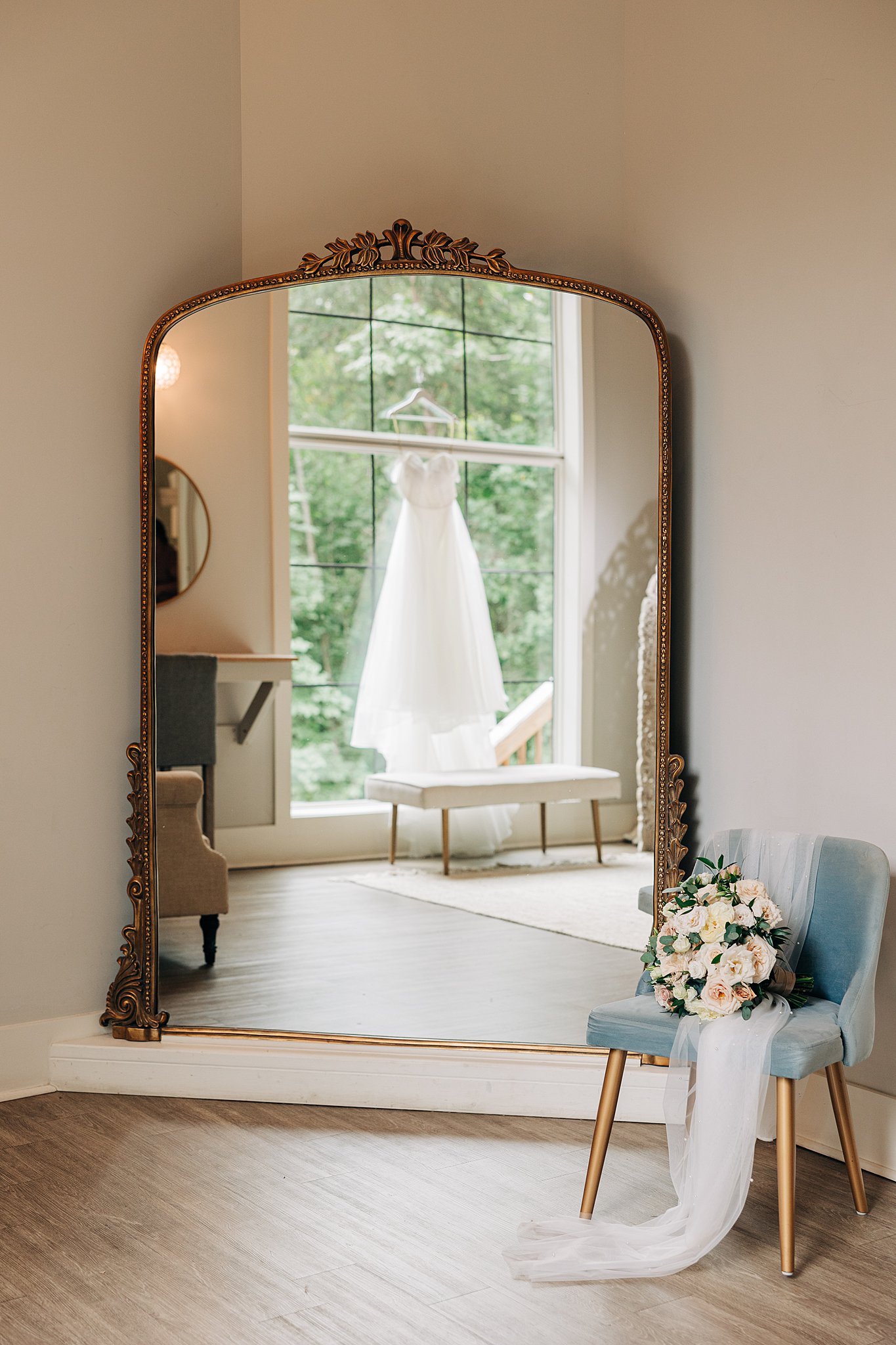 A brides dress hangs in a window in a mirror with a chair holding the veil and bouquet