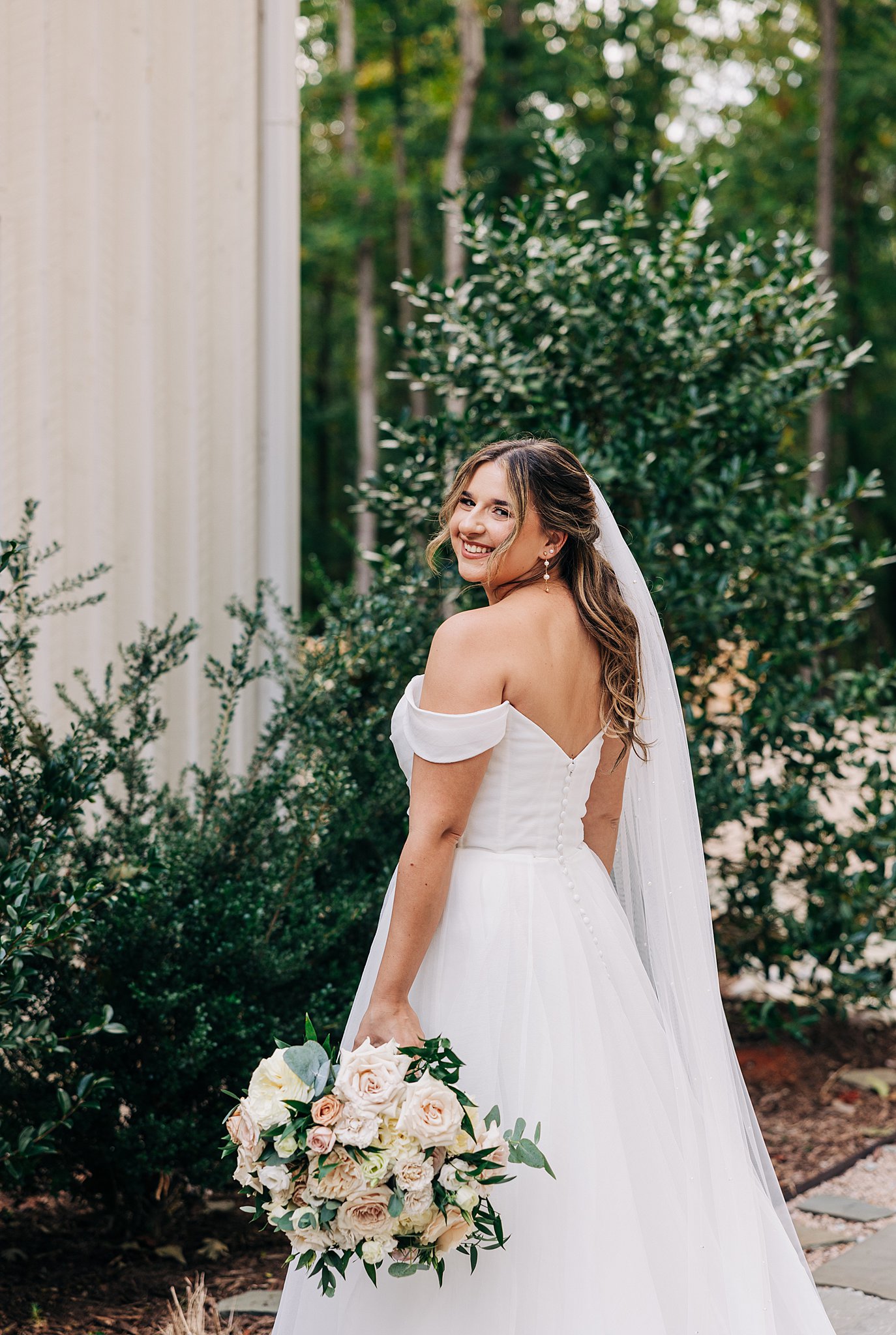 A bride in a silk dress smiles over her shoulder while standing in a garden