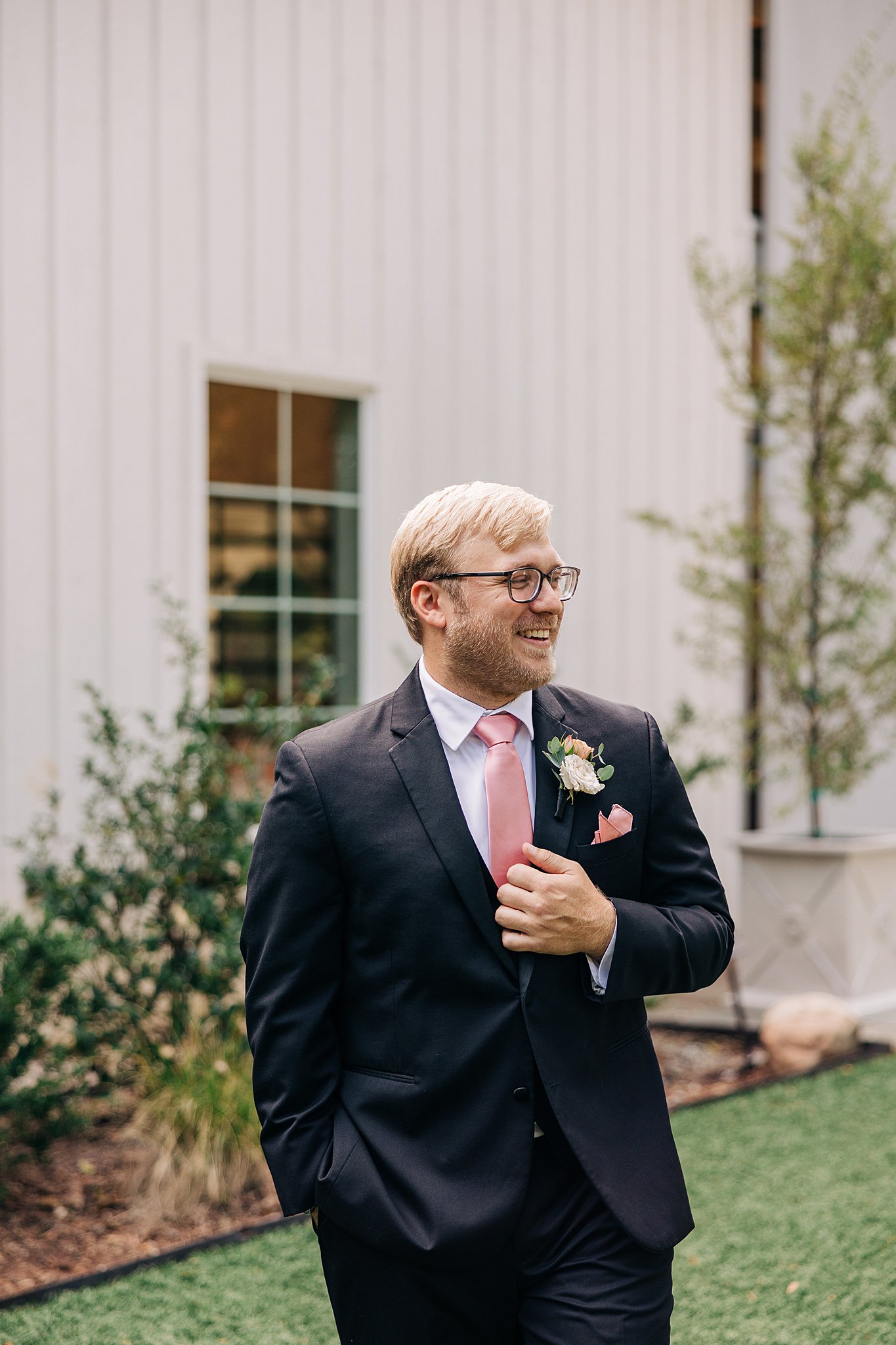 A groom laughs while standing in a garden in a black suit with pink accents