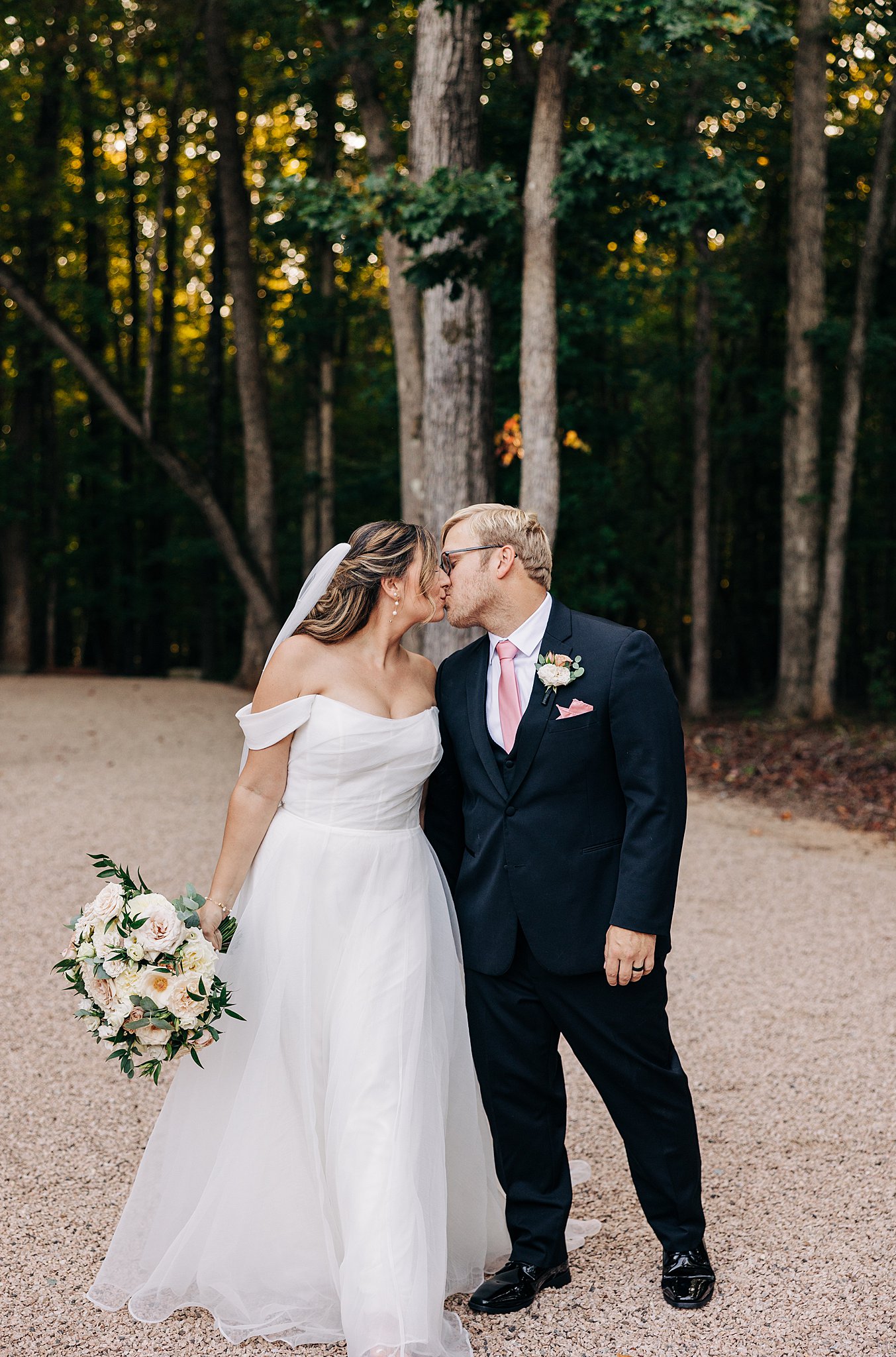 A bride and groom stand on a gravel path in the woods and share a kiss