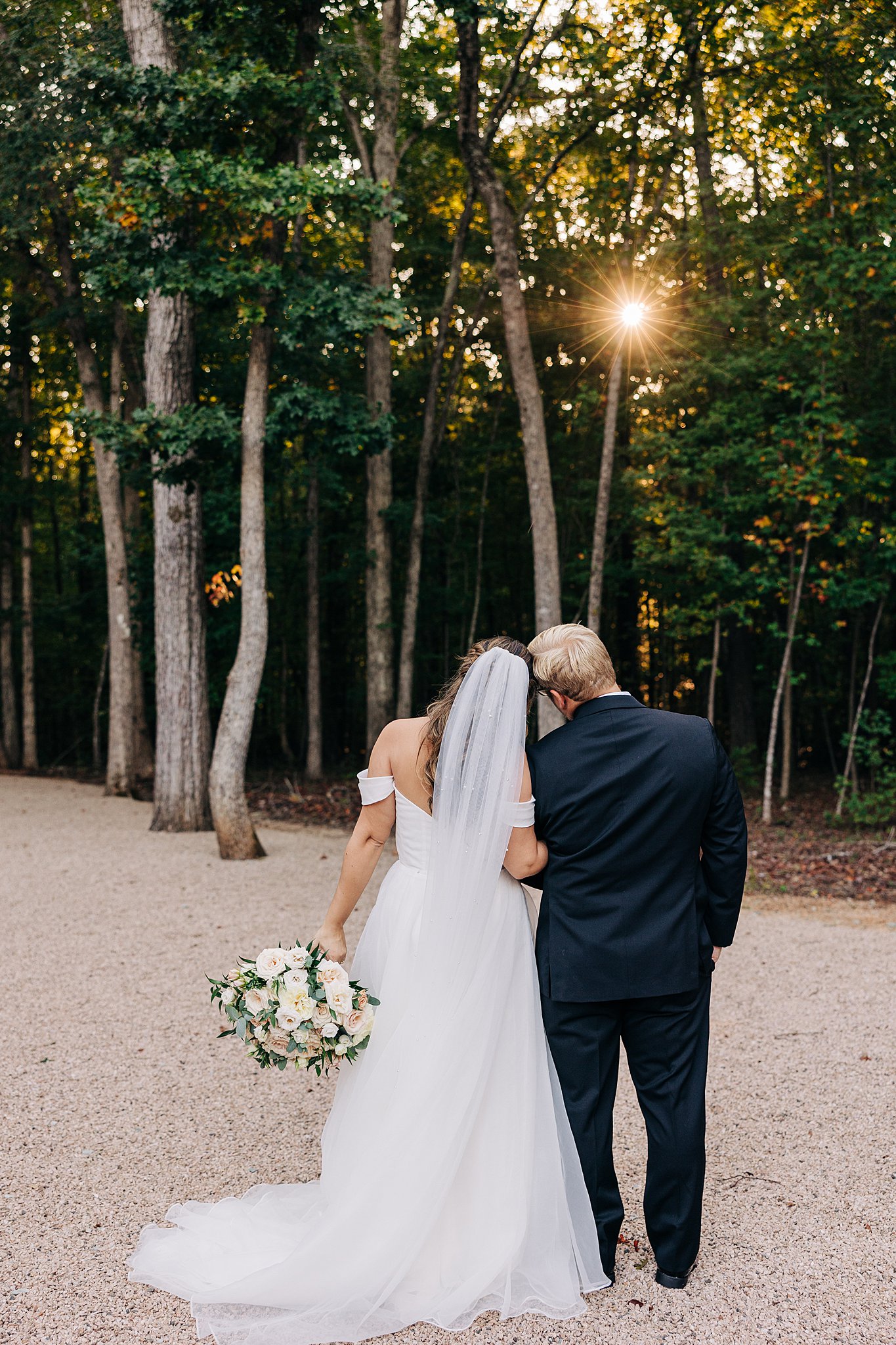 Newlyweds stand together touching heads while watching the sun set through the trees at their carolina grove wedding venue