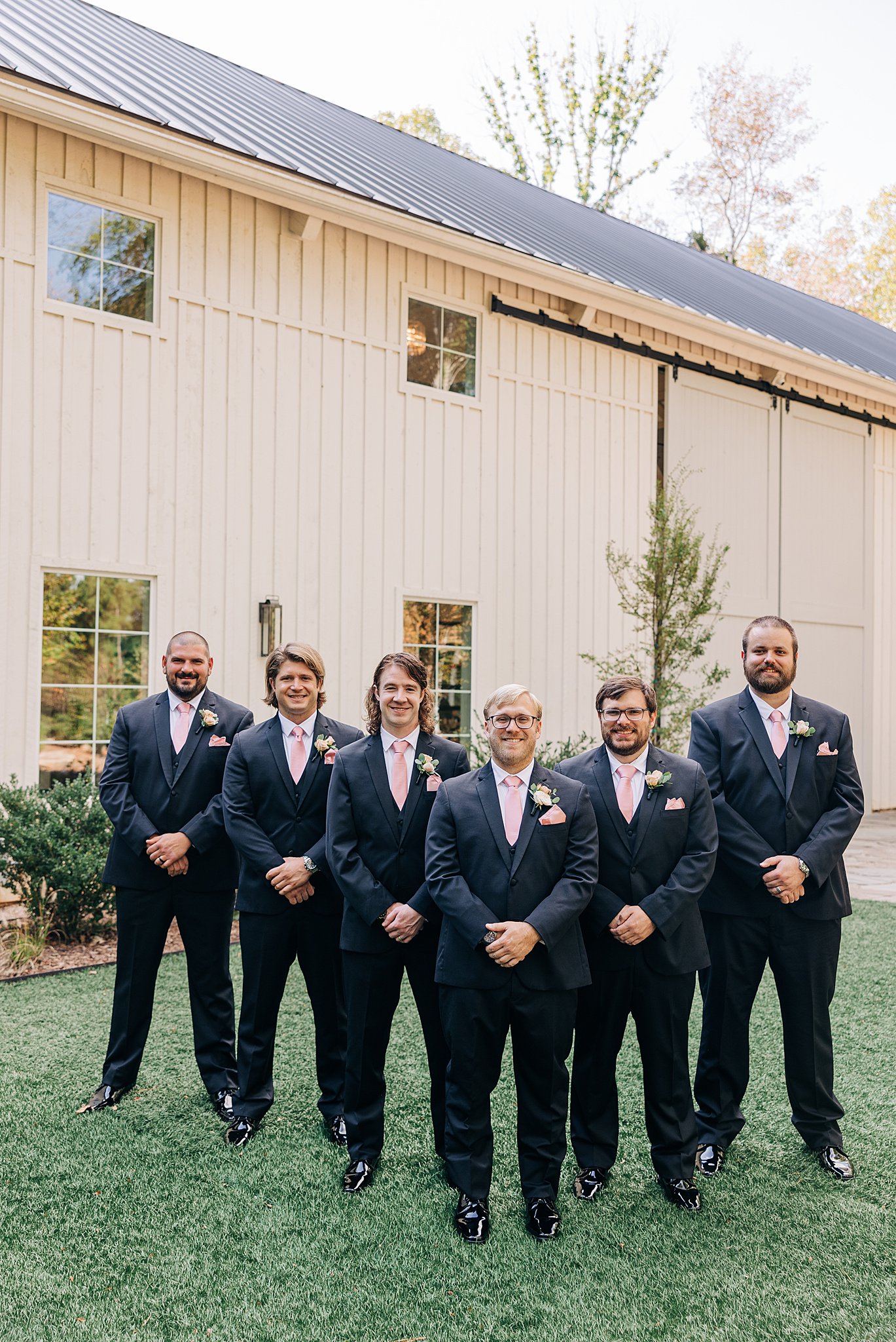 A groom in a black suit stands with his groomsmen in a garden lawn with hans folded in front