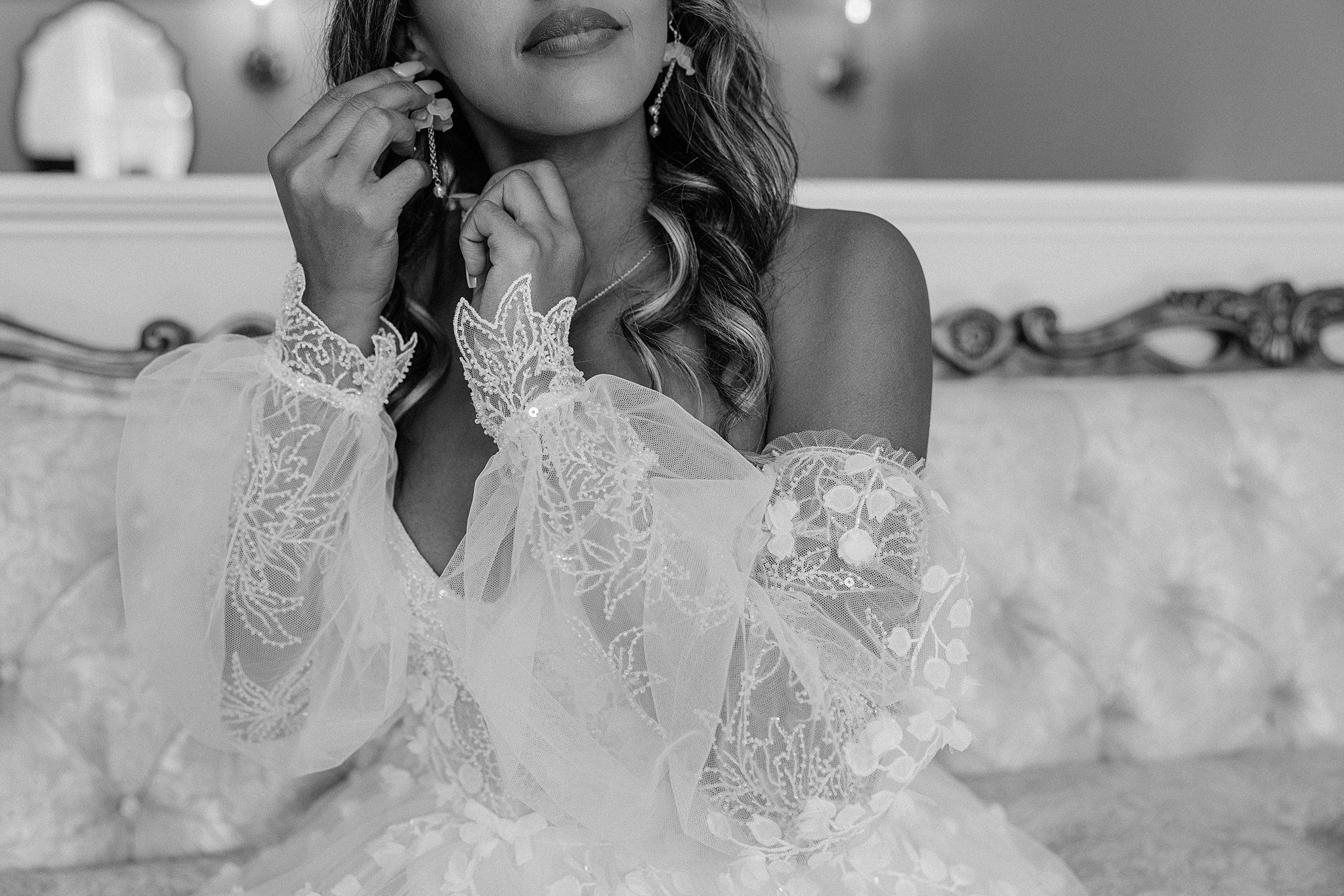 Details of a bride in a lace dress putting on her earrings on an antique couch