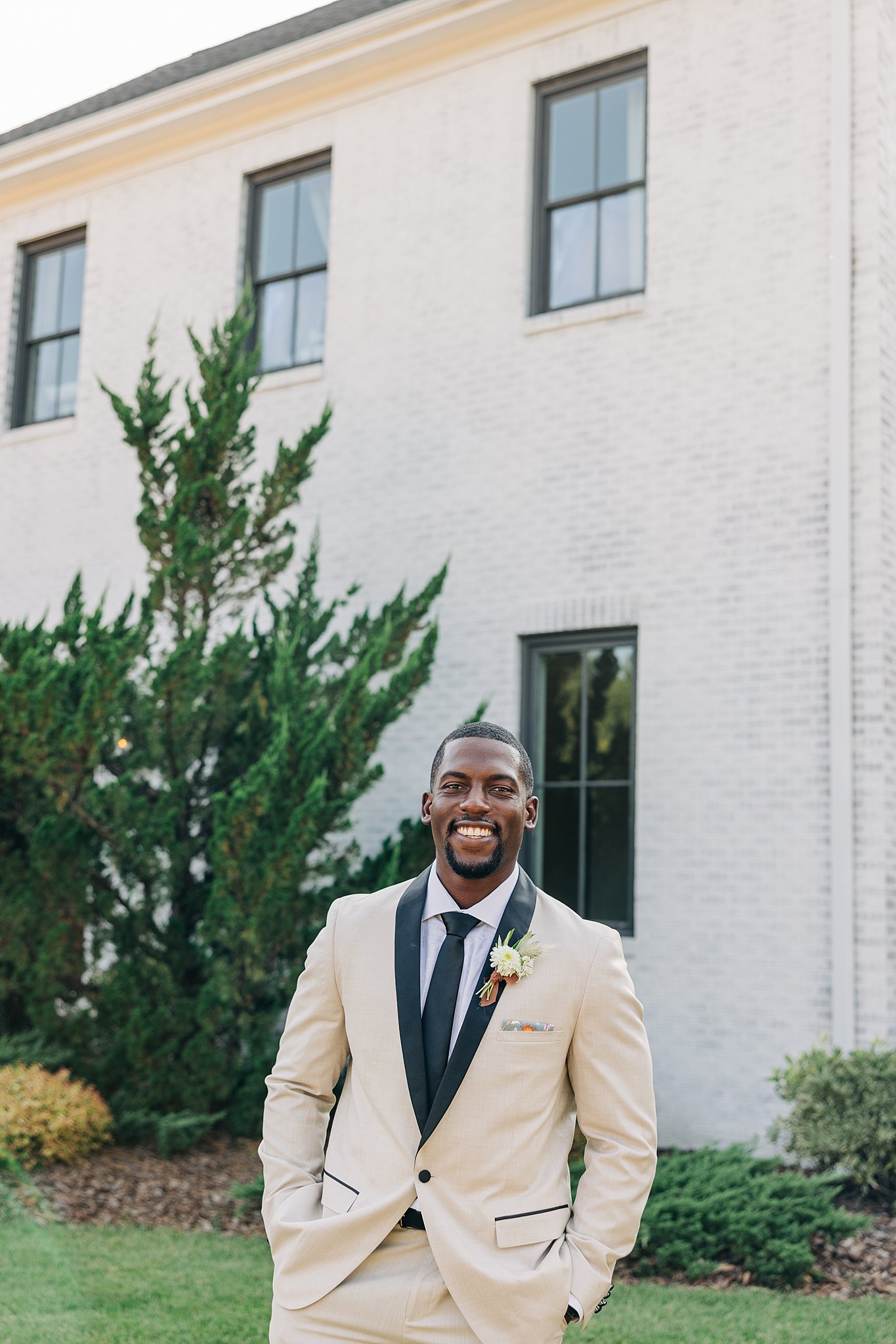 A groom in a cream and black suit stands in a back garden smiling with hands in pockets