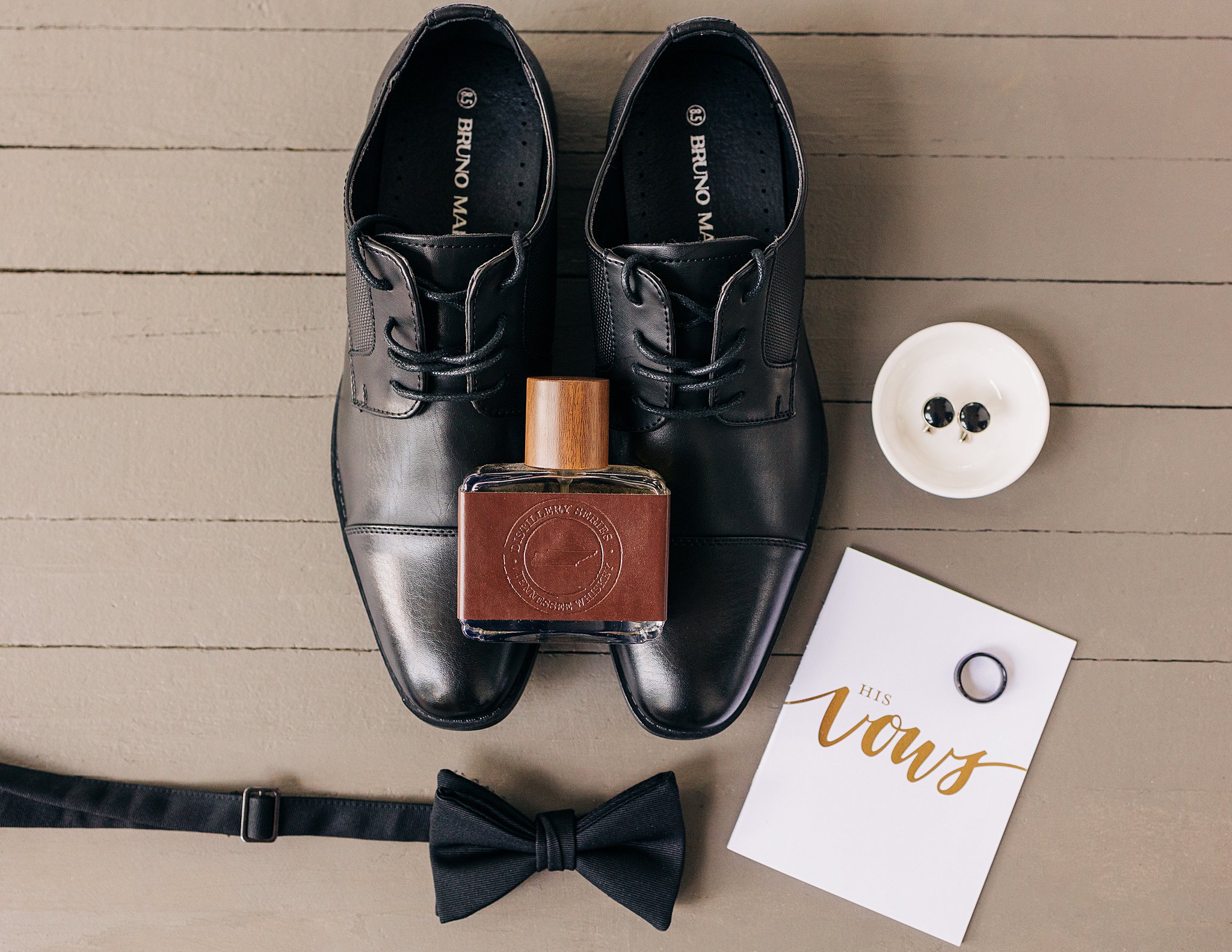 Groom details on a wooden floor with black shoes, cologne, bowtie and vows