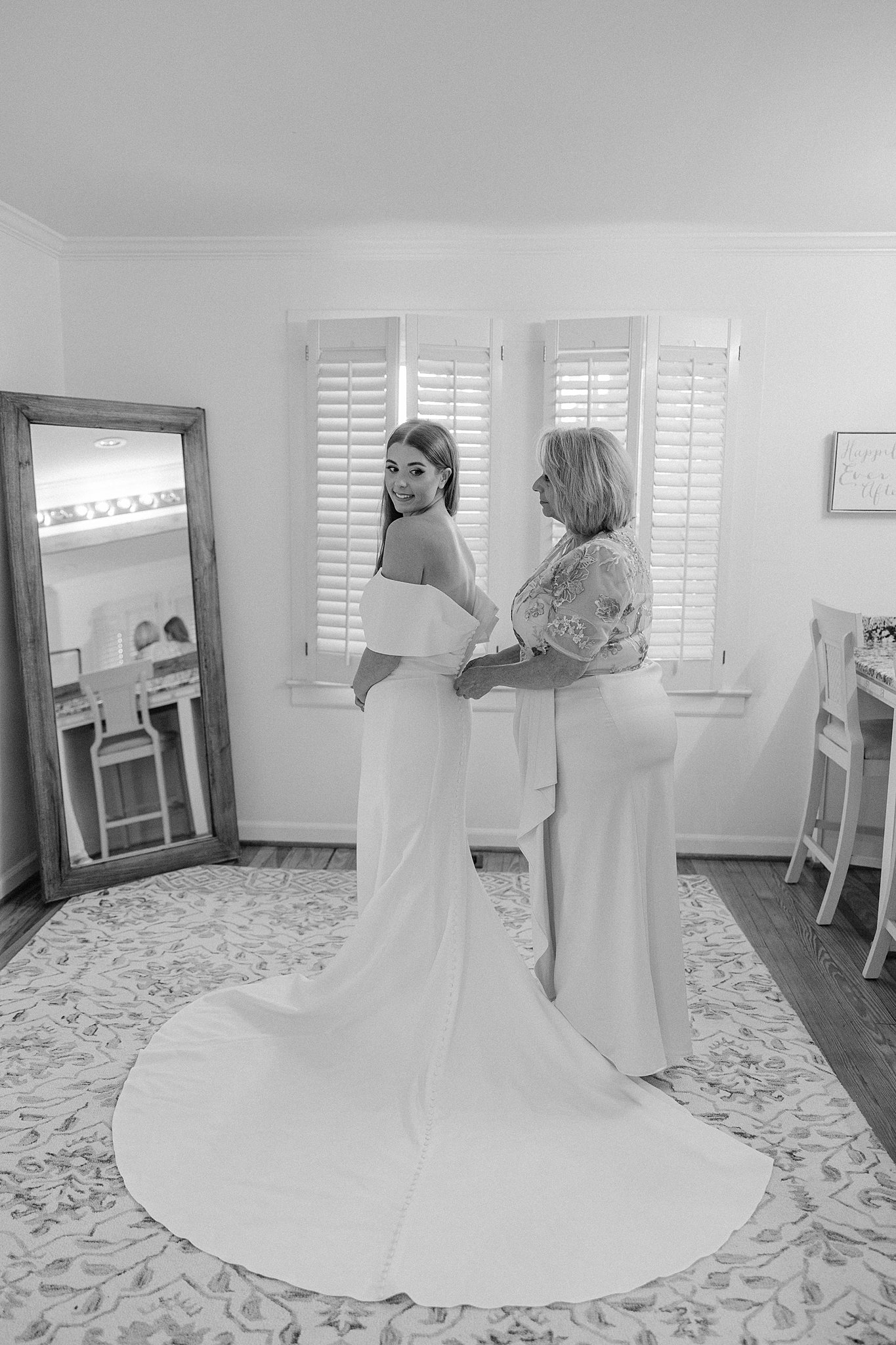 A mother helps zip up her daughter's wedding dress while standing in front of a mirror