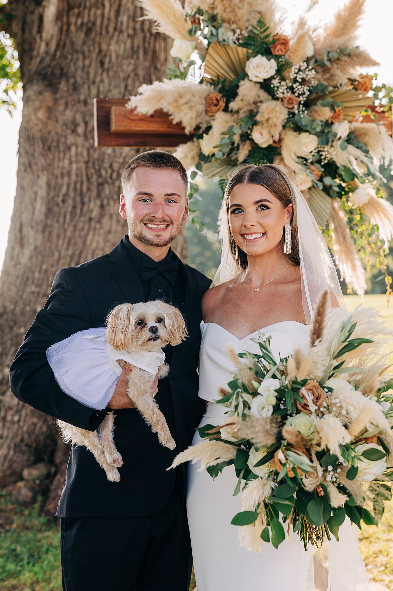 A groom in a black suit holds a dog in a white dress while standing with his bride holding her large bouquet