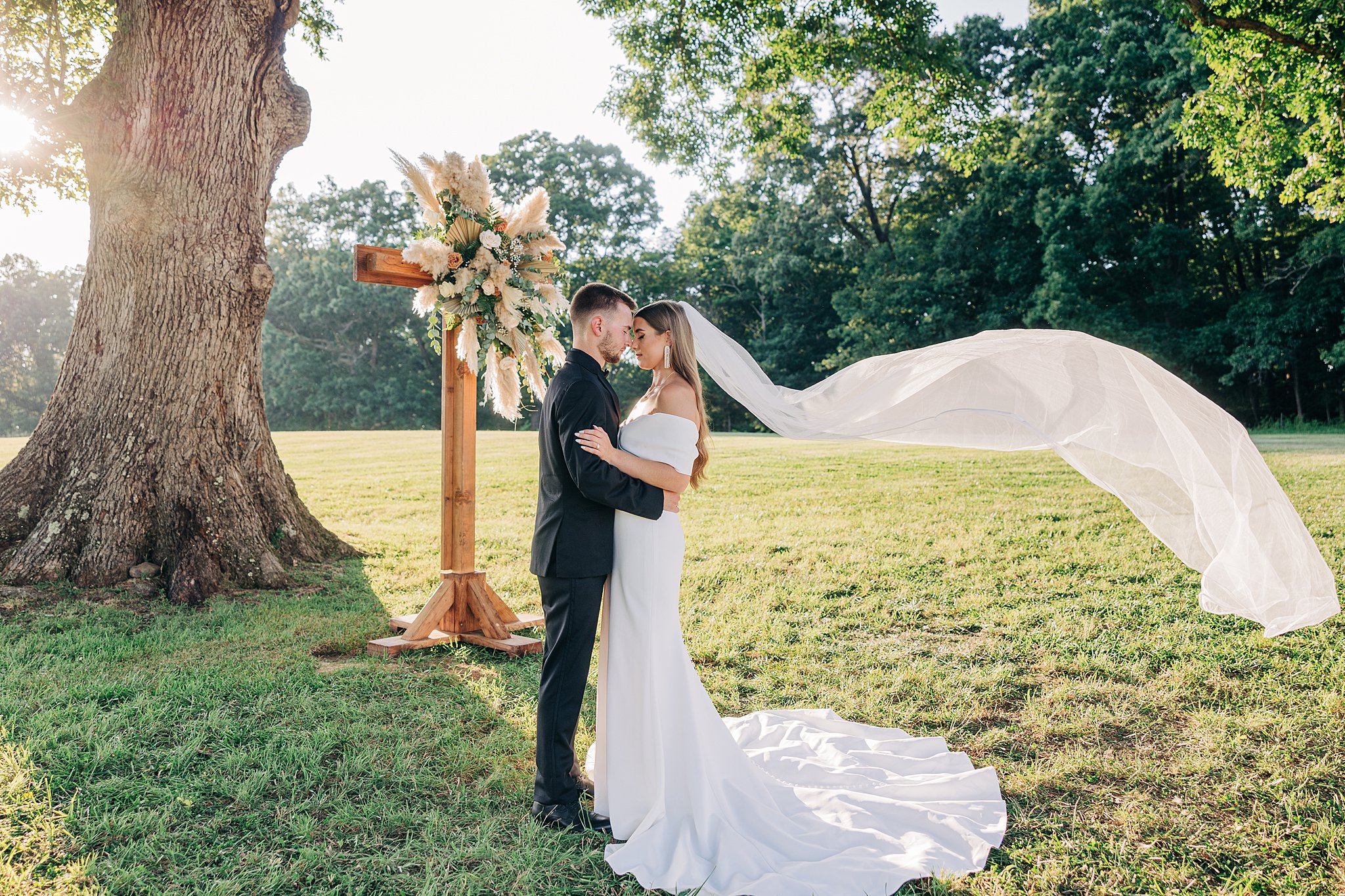Newlyweds stand close touching foreheads in a windy field under an oak tree with a wooden flower covered cross at their Summerfield Farms wedding