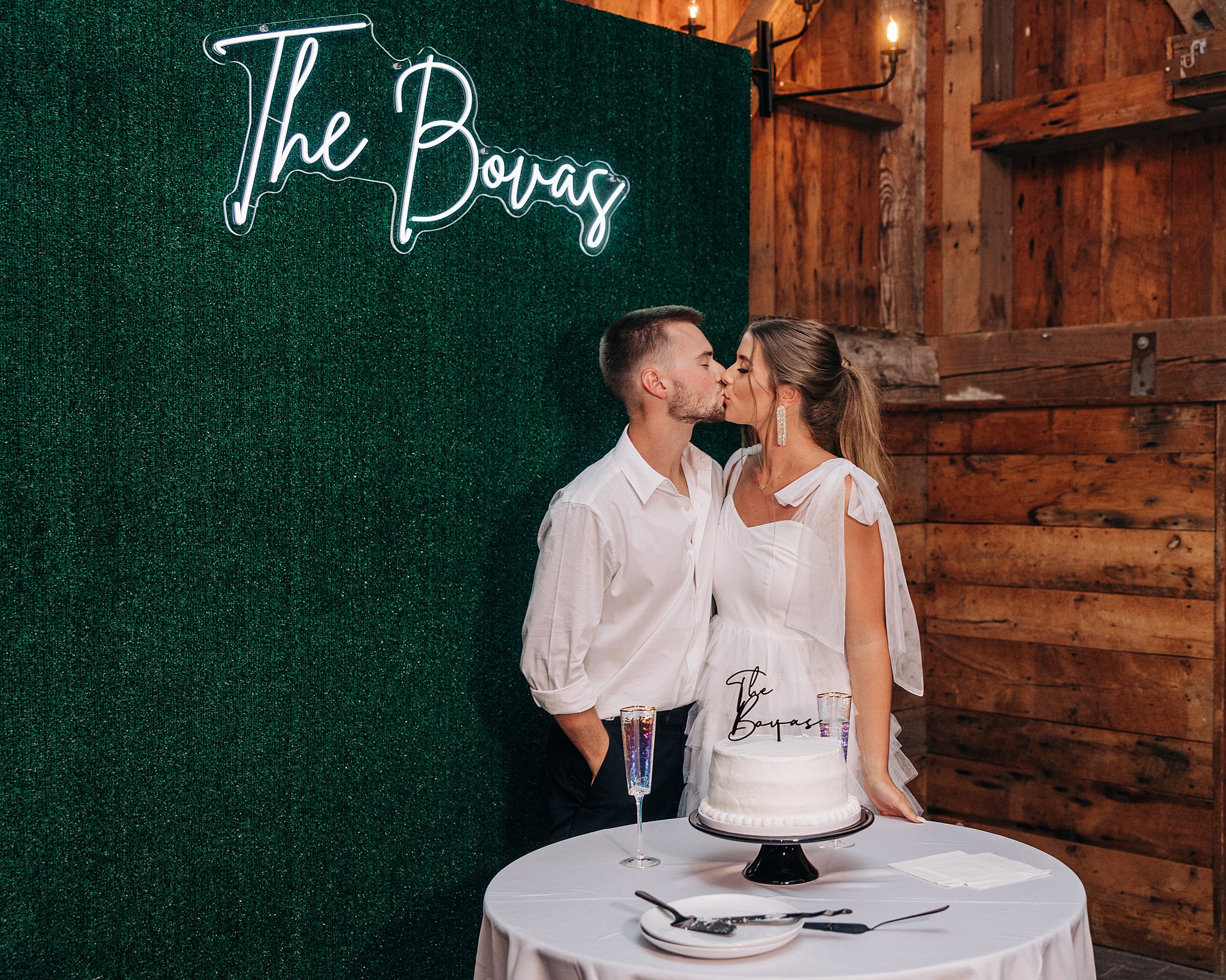 Newlyweds kiss while standing behind their white cake in a barn