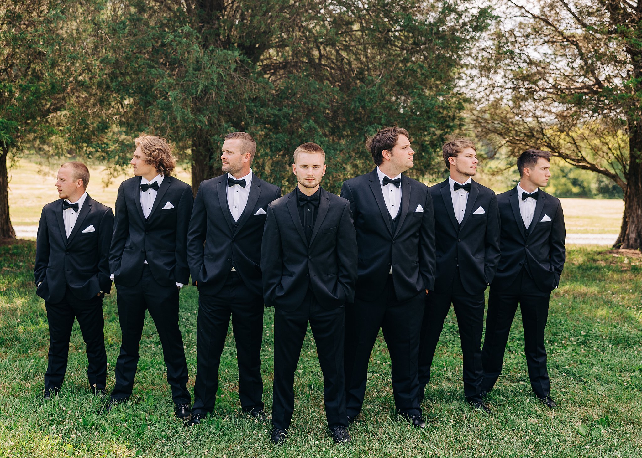 A groom stands with his hands in his pockets in a black suit with his groomsmen around him