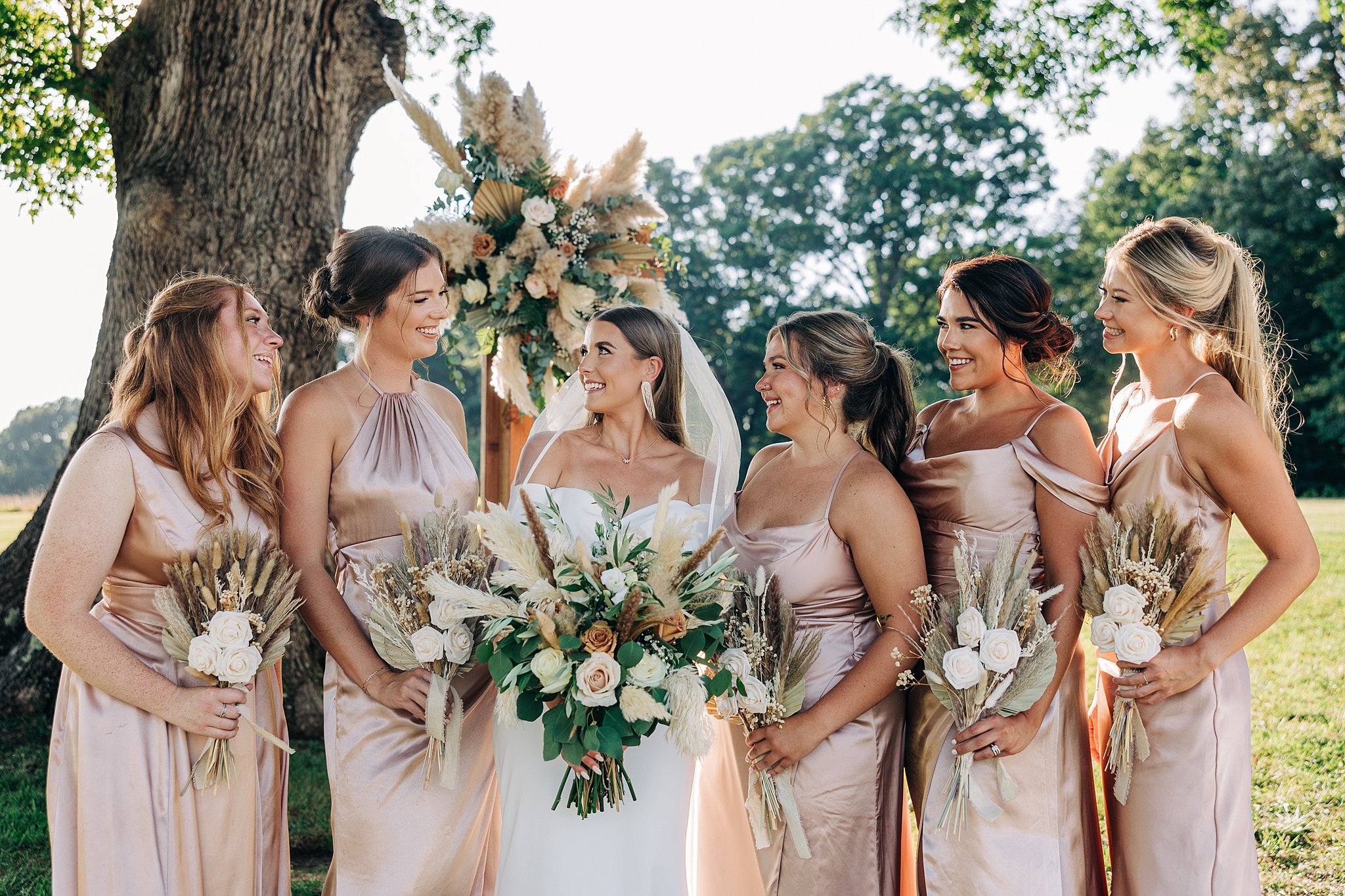 A bride smiles while standing with her bridal party under a large oak tree at her Summerfield Farms wedding