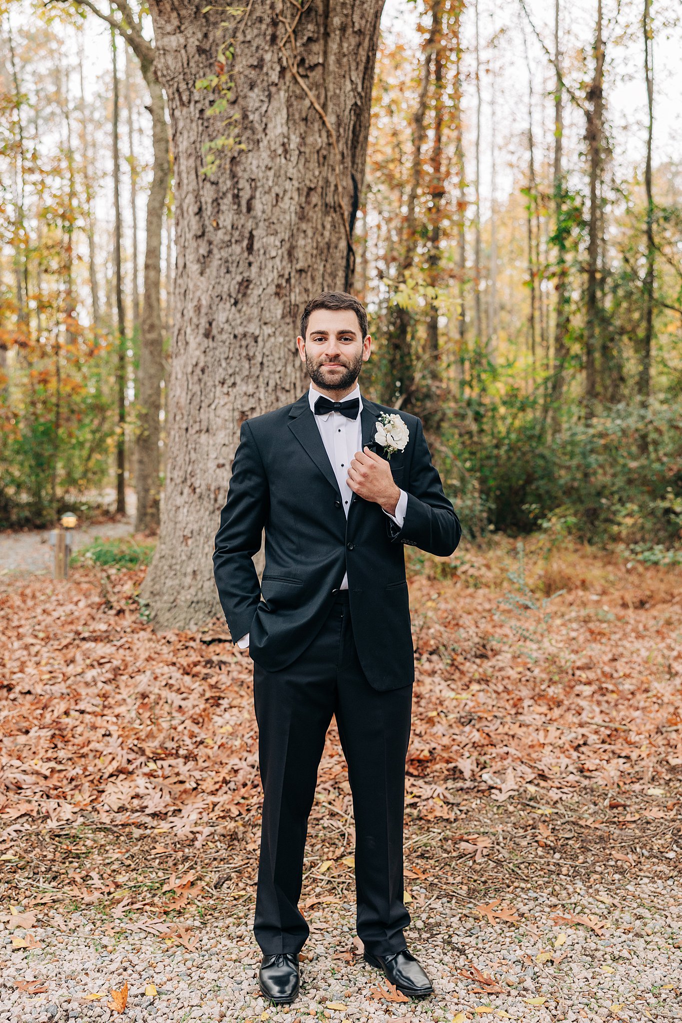 A groom holds onto his lapel while standing under a tall old tree in fall
