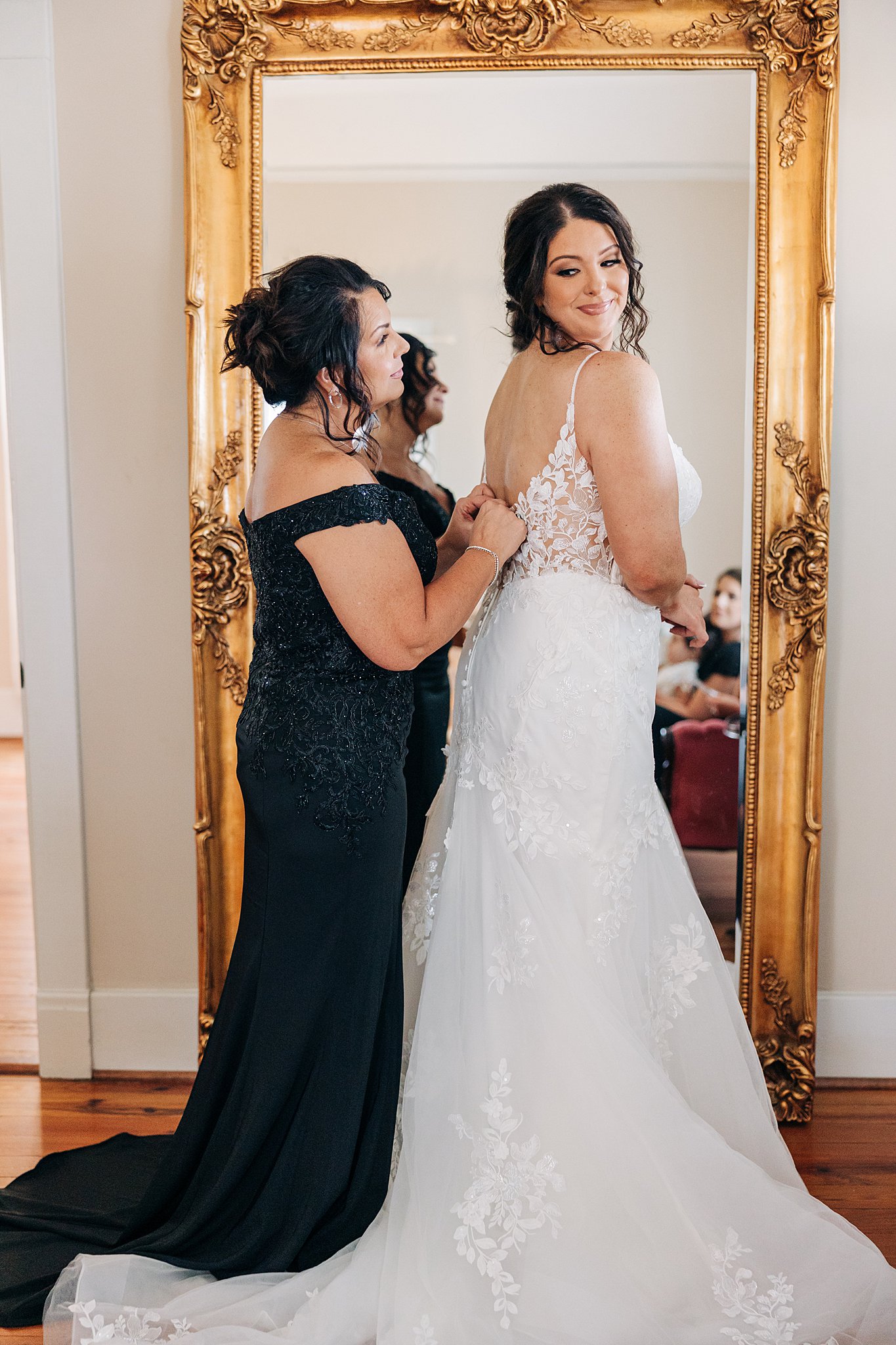 A bride has help buttoning her dress while standing in a mirror