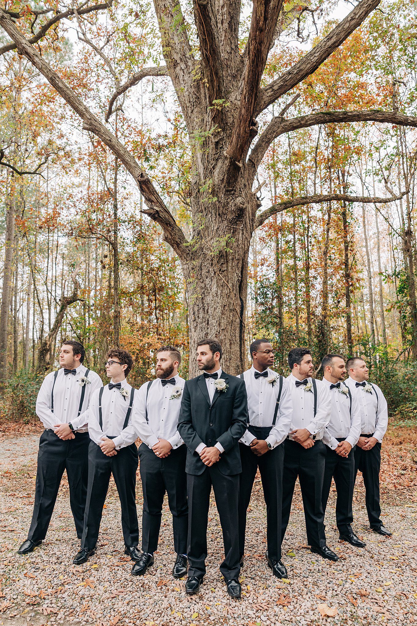 A groom stands under a large tree with hands in front of him with his seven groomsmen