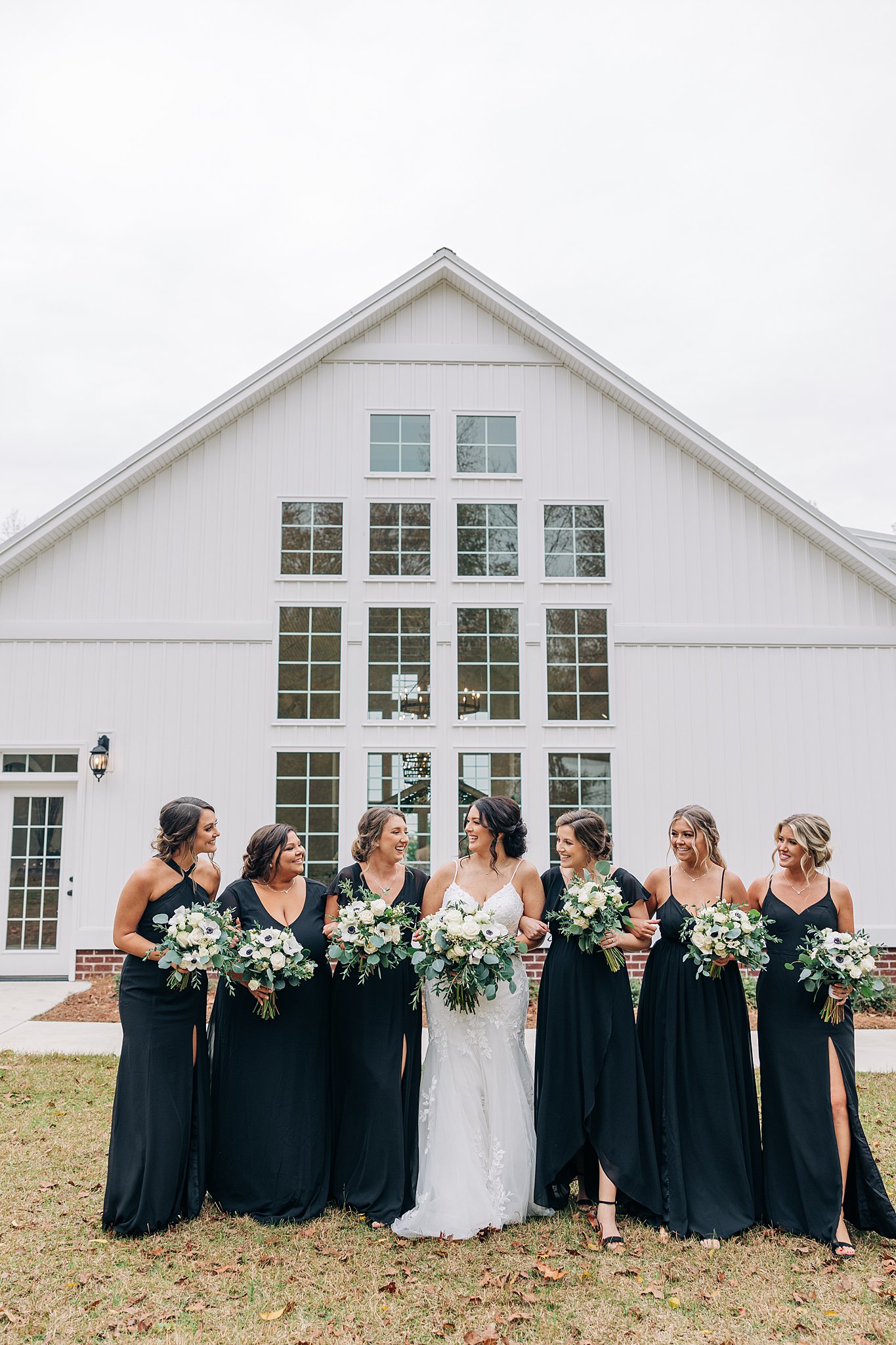 A bride stands in a lawn laughing with her six bridesmaids in black dresses and holding bouquets