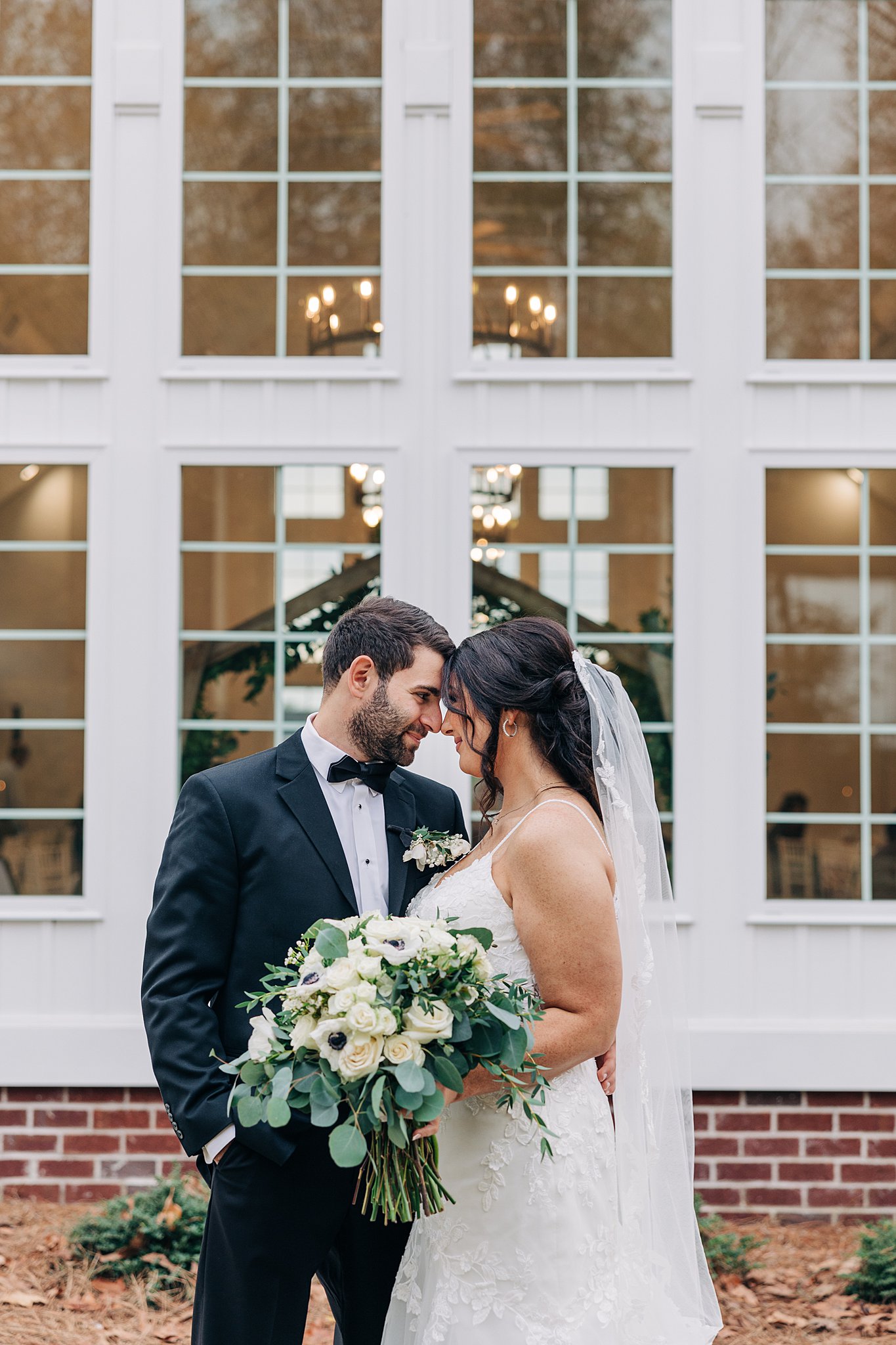 Newlyweds touch foreheads while sharing a quiet moment in a garden in a black tux and white lace dress