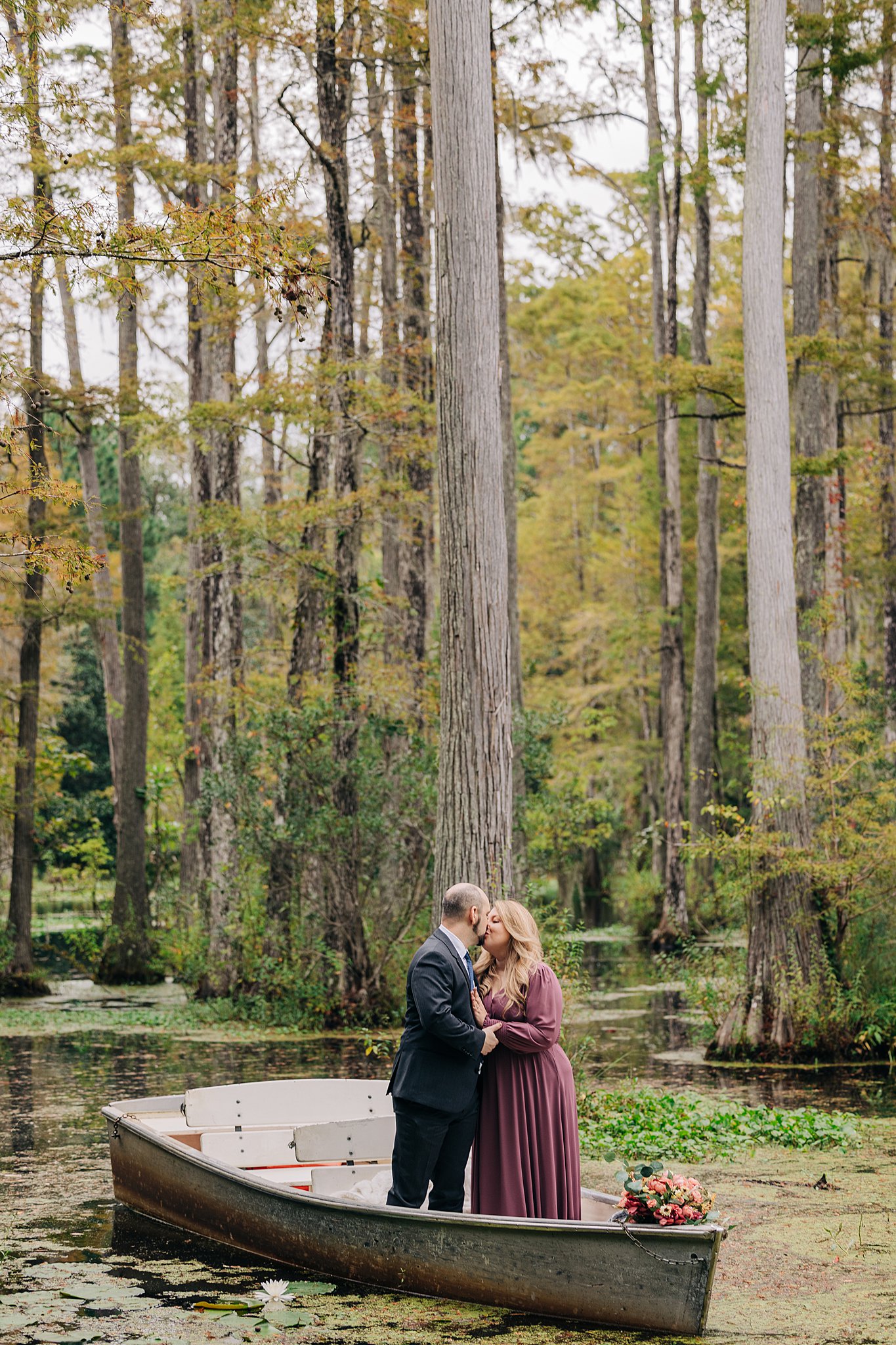 A newly engaged couple in a dark suit and pink dress kiss while standing in a boat in the cypress gardens wedding engagement venue
