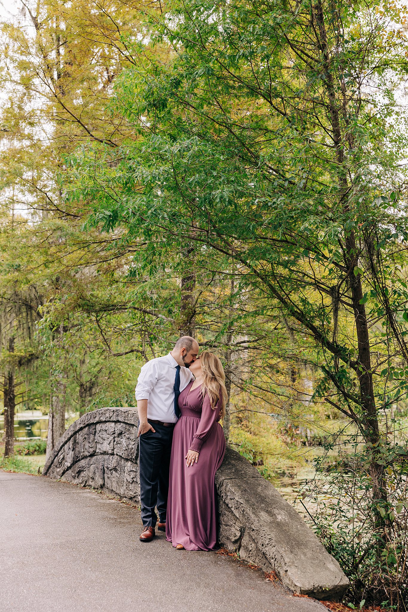 A couple kisses while sitting on a stone bridge railing during their cypress gardens wedding engagement