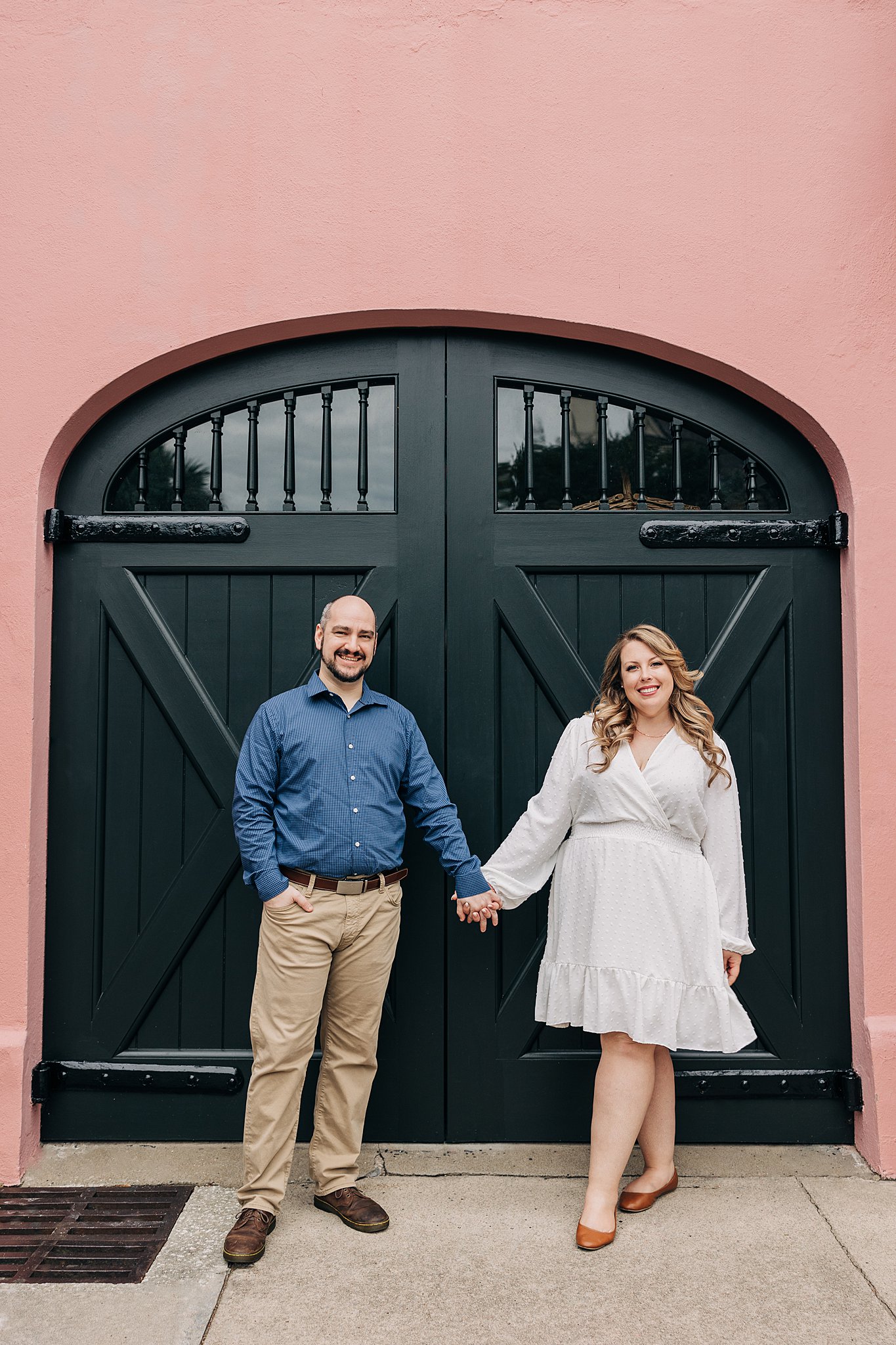 A newly engaged couple hold hands in a large wooden doorway of a pink building