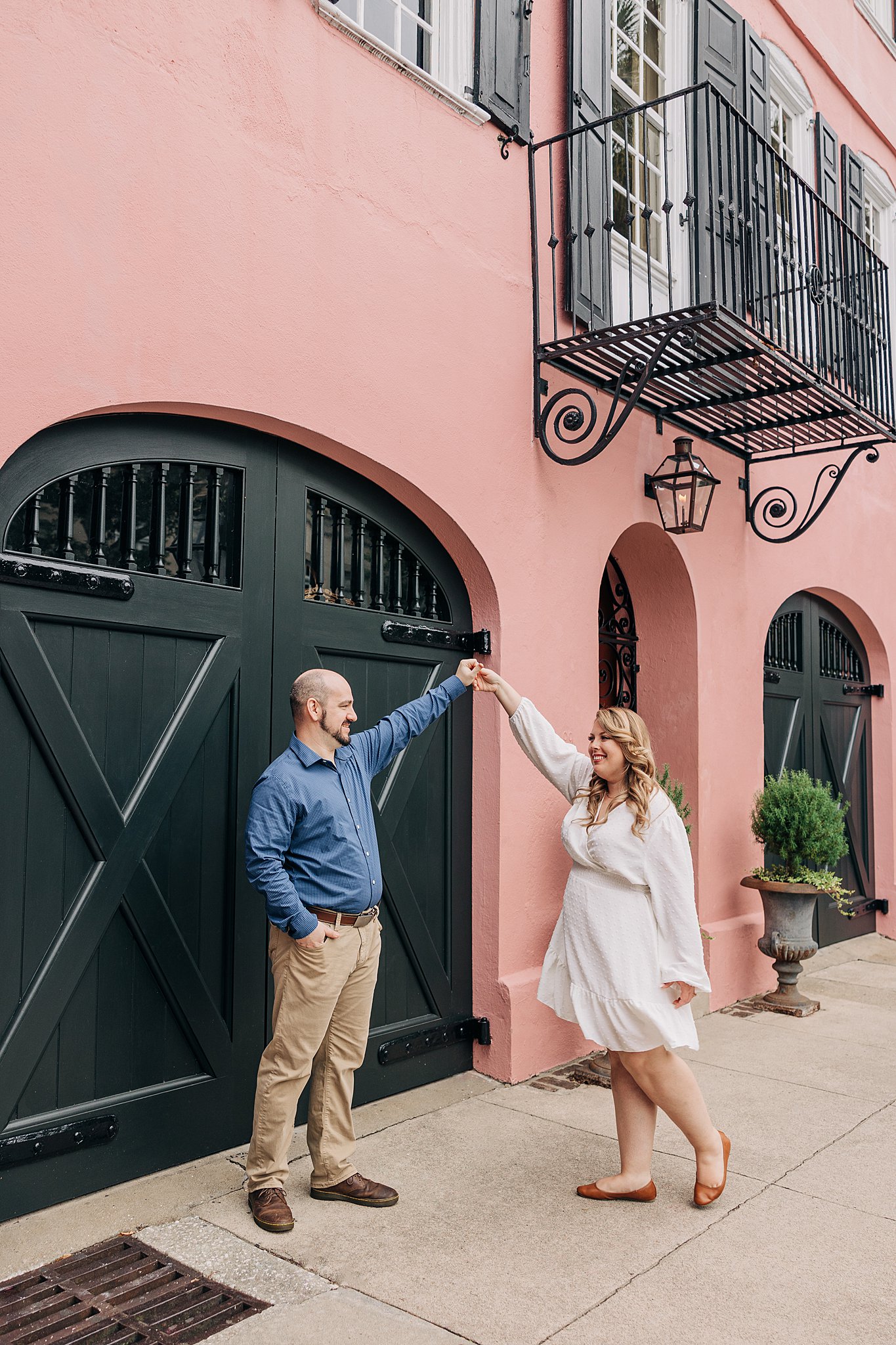 A newly engaged couple dance in an alley by a pink building in front of large black wooden doors