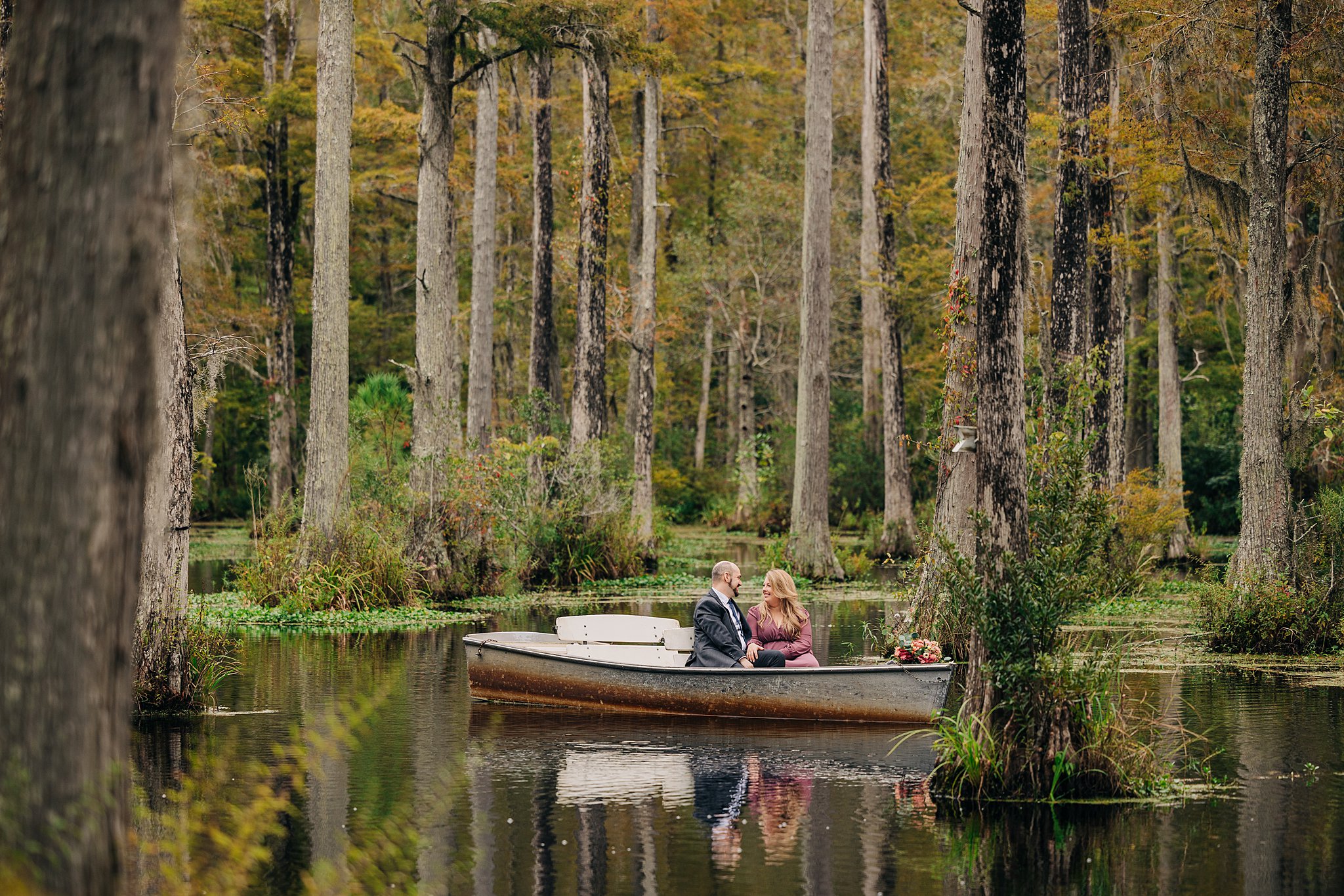 A couple smiles at each other in a pink dress and blue suit while sitting in a small boat during their cypress gardens wedding engagement