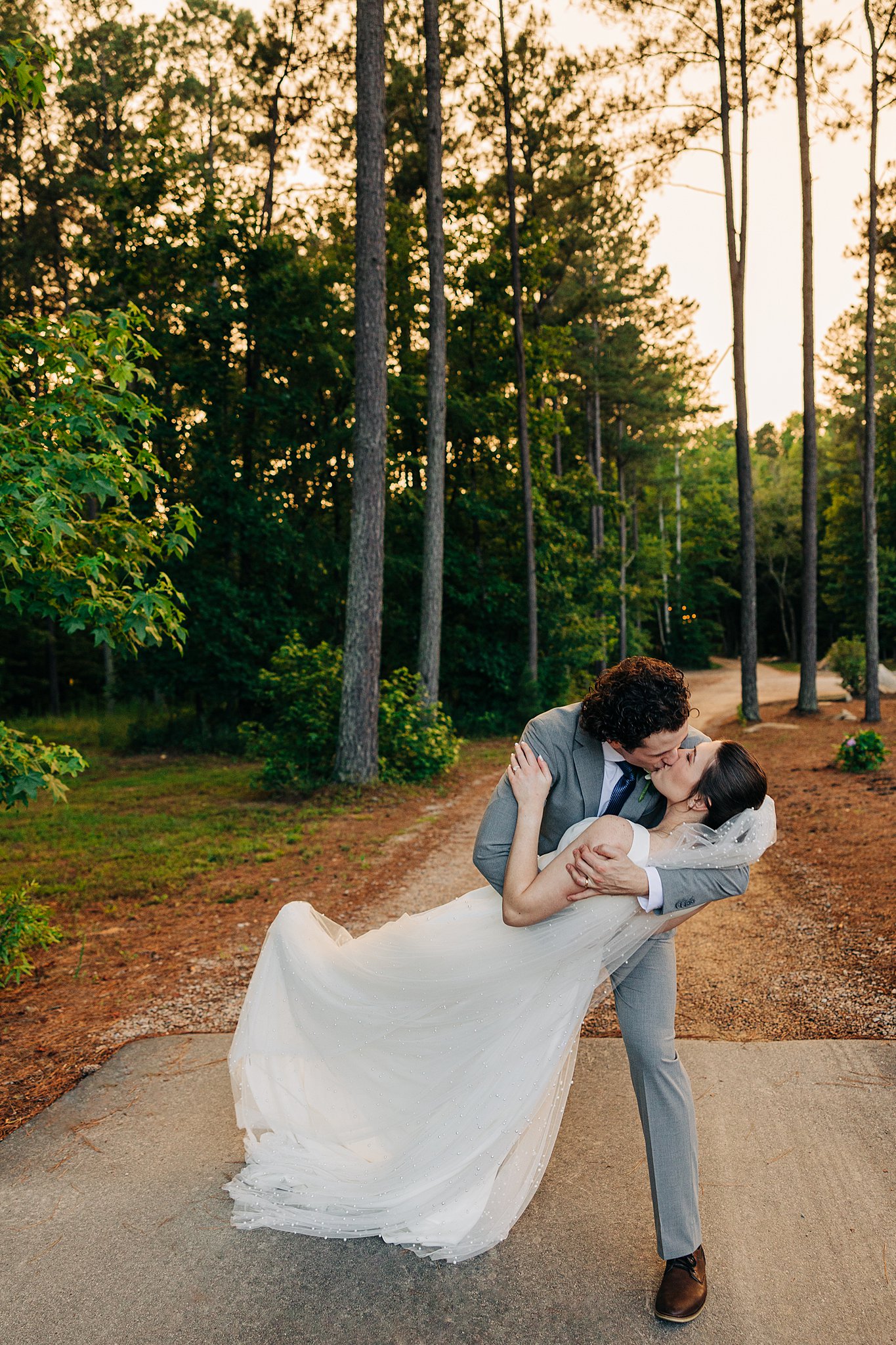 A groom in a grey suit dips his bride in a forest path at sunset at the pinehill pavilion