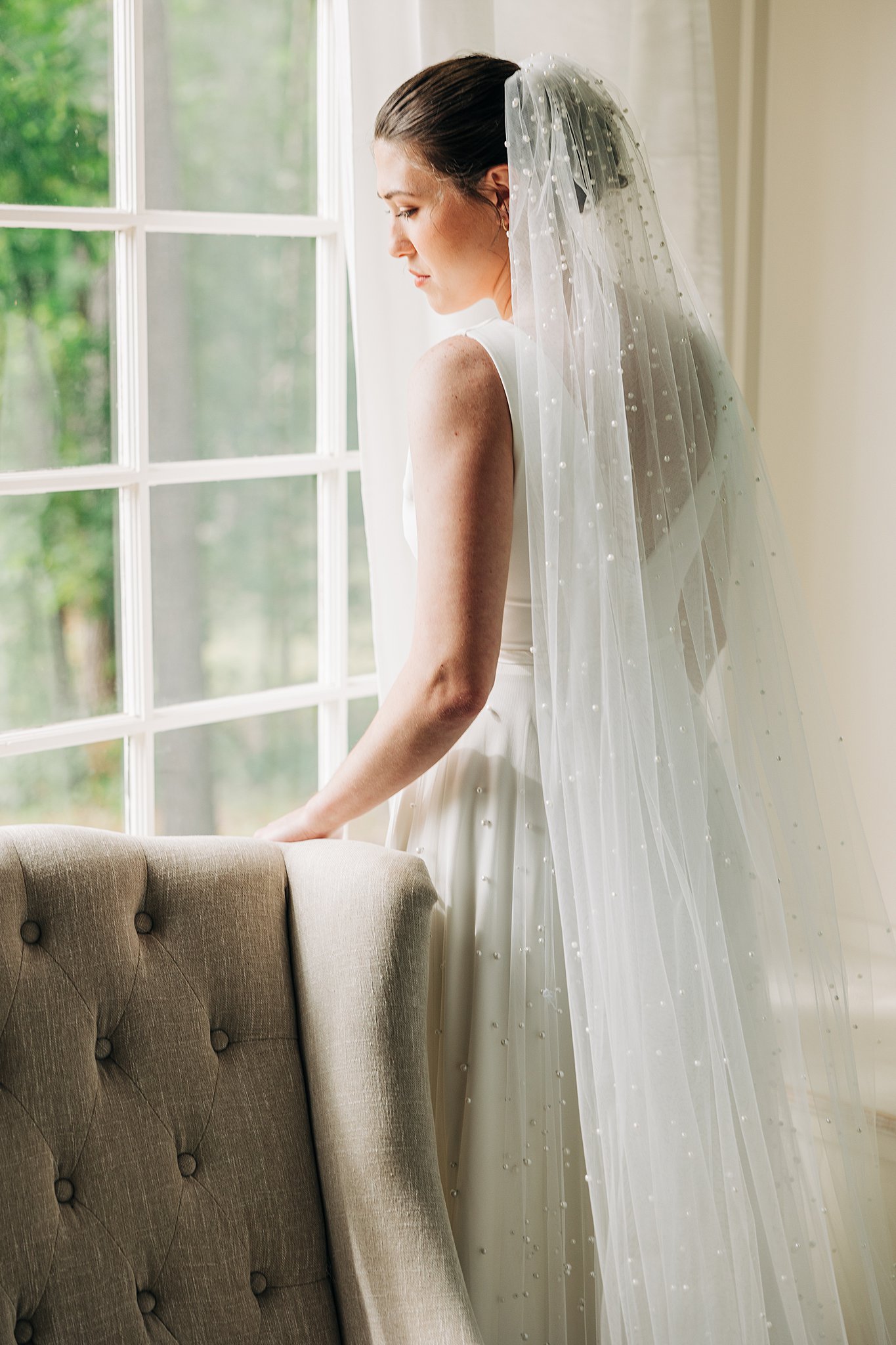 A bride gazes out a window while leaning against a chair before her pinehill pavilion wedding