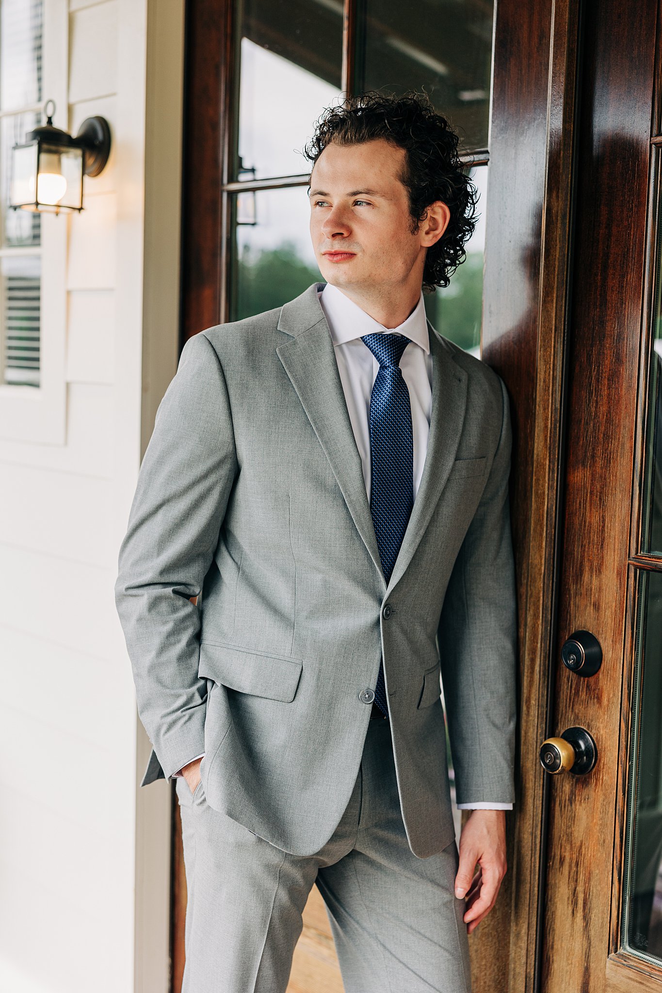 A groom in a grey suit leans against a wooden door on a porch
