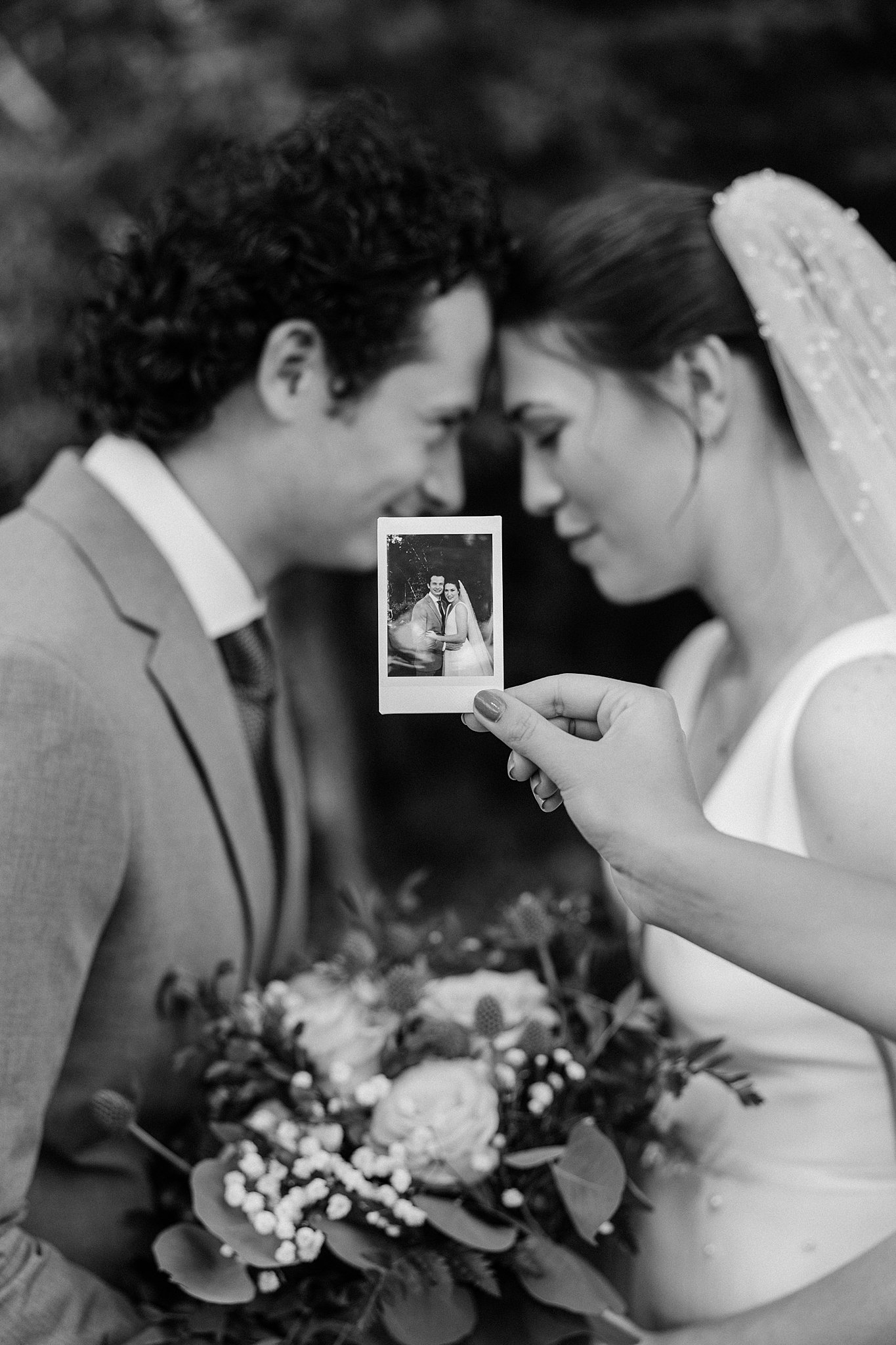 A polaroid is held in front of a newlywed couple touching foreheads of them