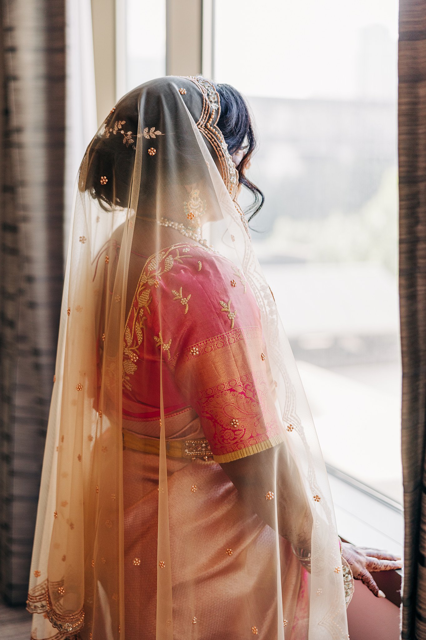 A bride in a pink traditional wedding outfit stands in a window