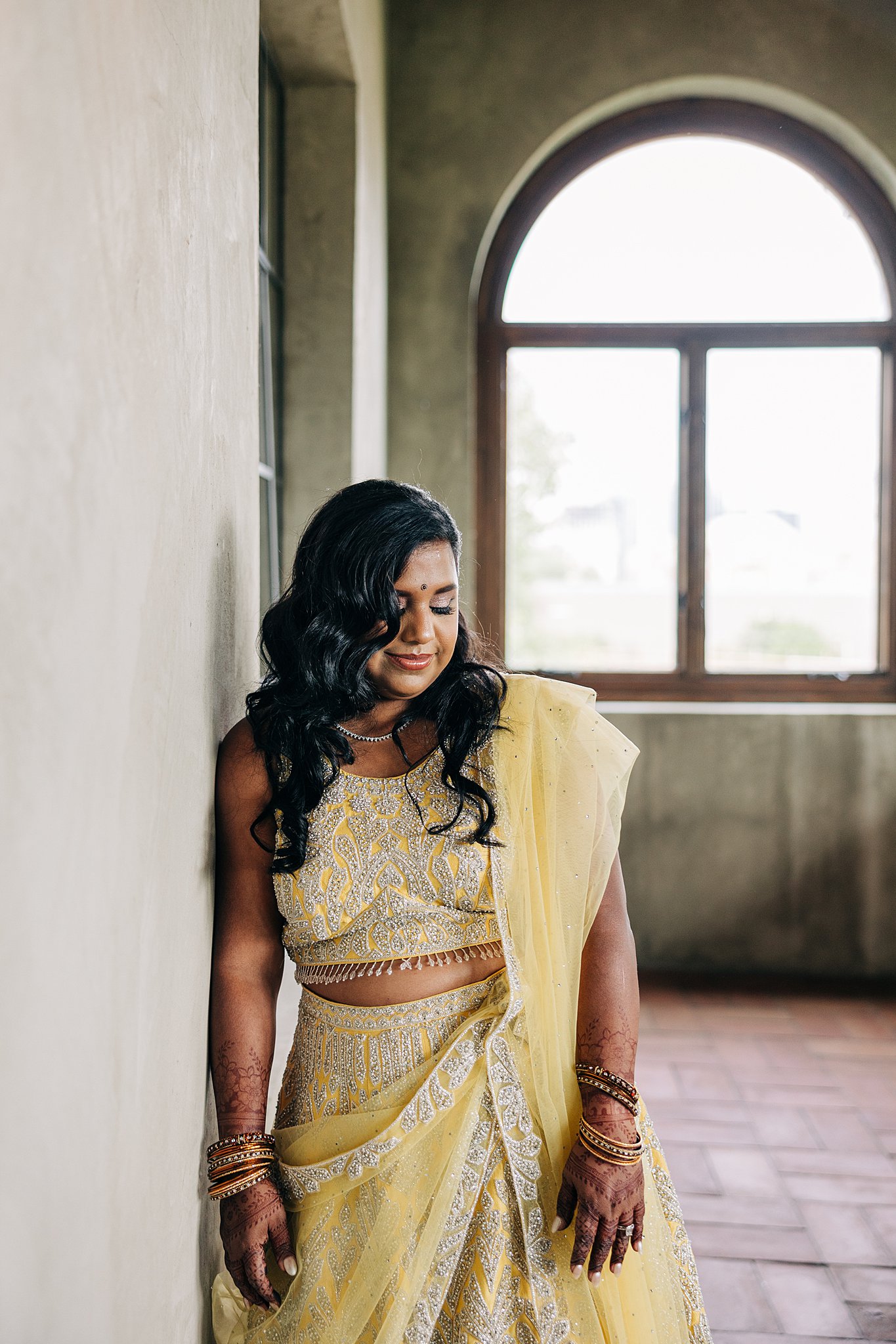 A bride in a traditional yellow dress leans against a wall smiling down her shoulder
