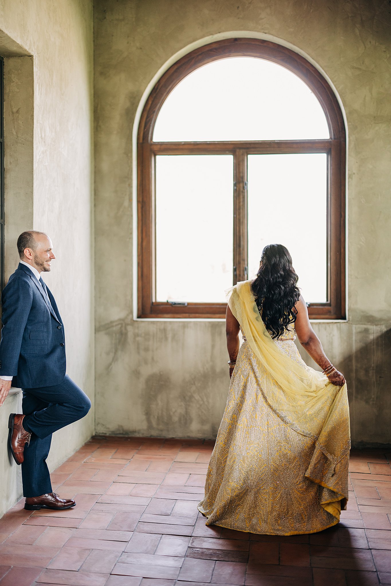 A groom leans on a wall in a blue suit while smiling at his bride in a yellow dress during their summerour studio wedding