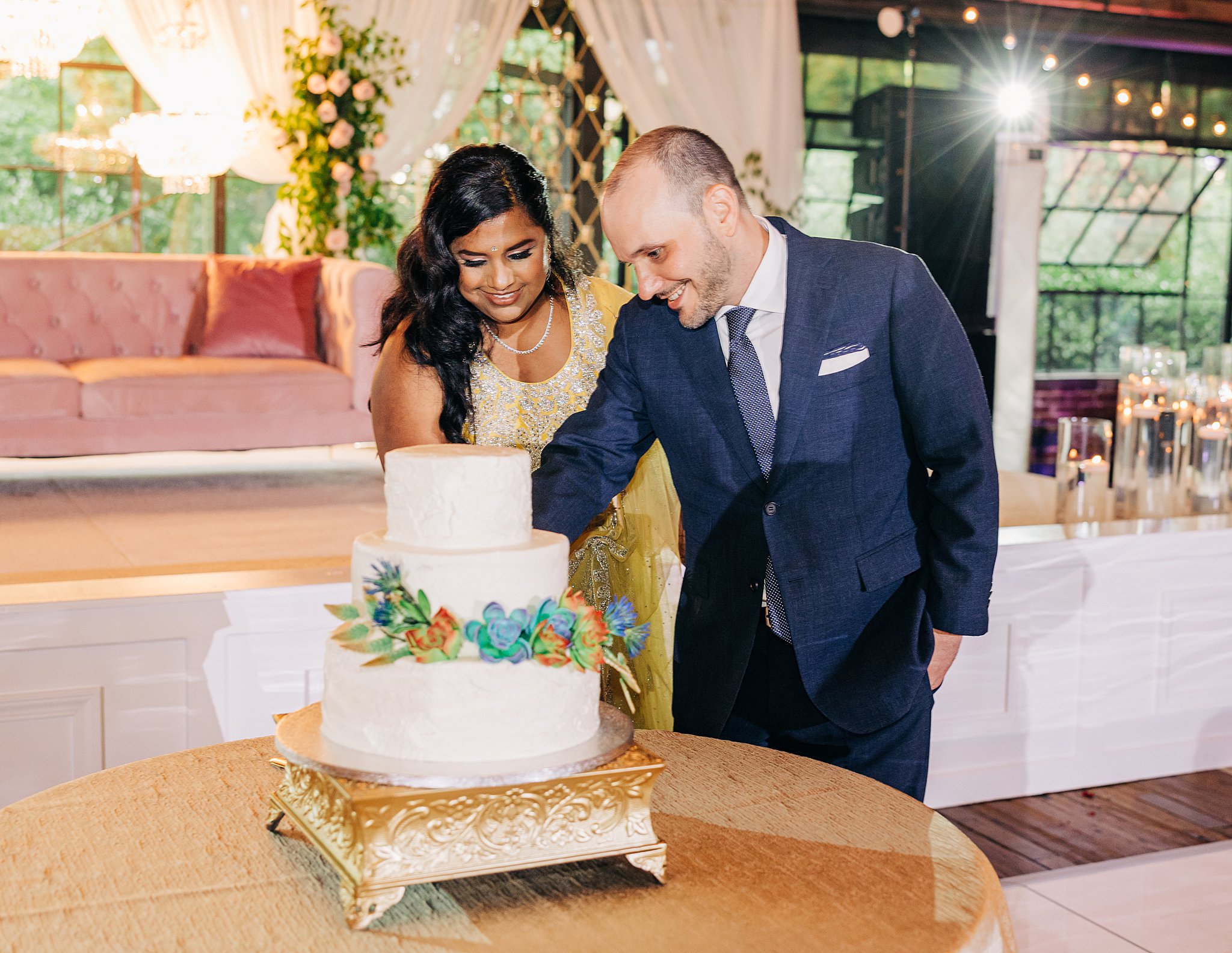 Newlyweds cut their white three-tier cake with colorful flowers in a blue suit and yellow traditional dress