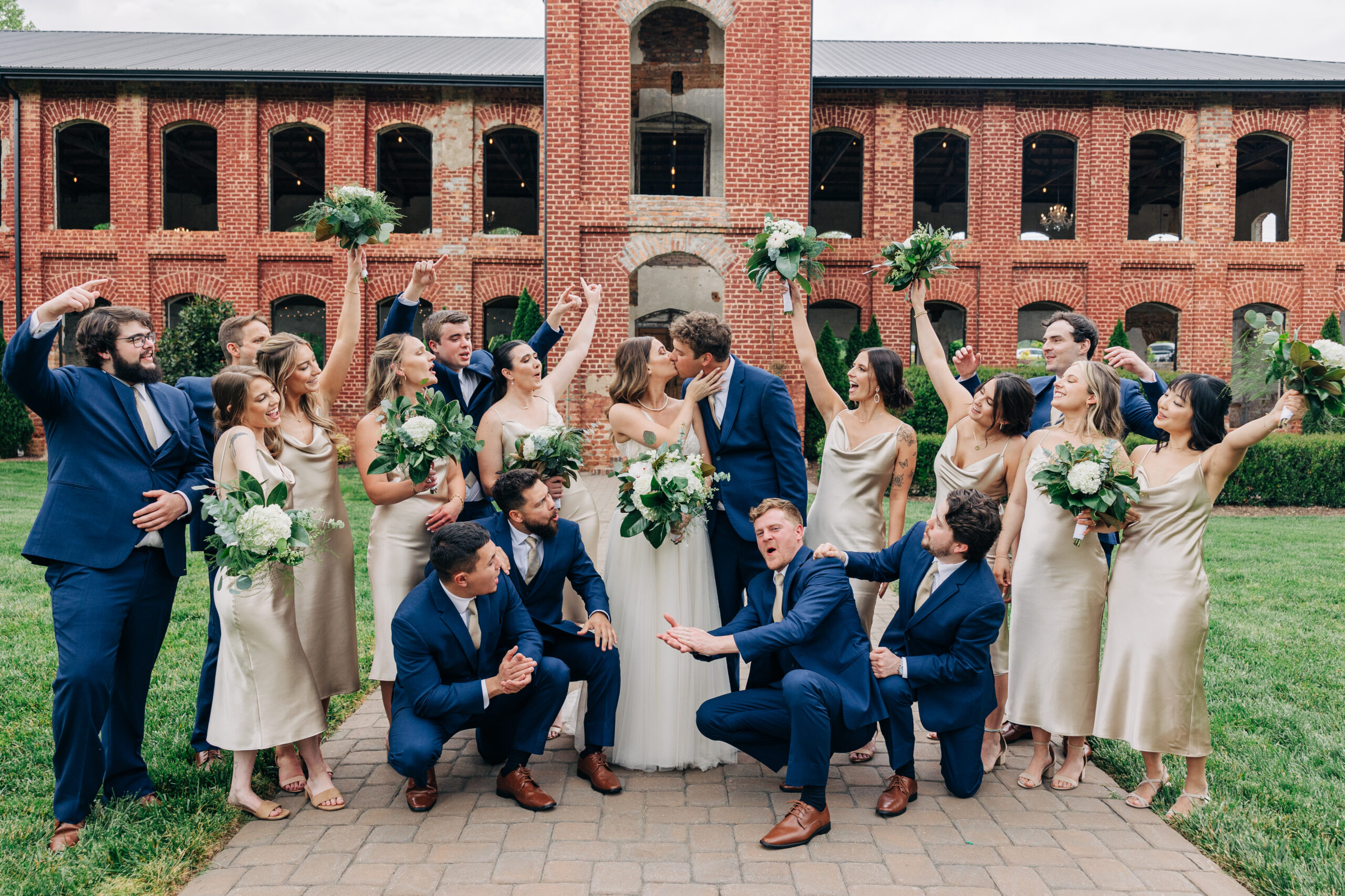 Newlyweds kiss while their large wedding party celebrate and cheer in the front walkway of the Providence Cotton Mill wedding venue