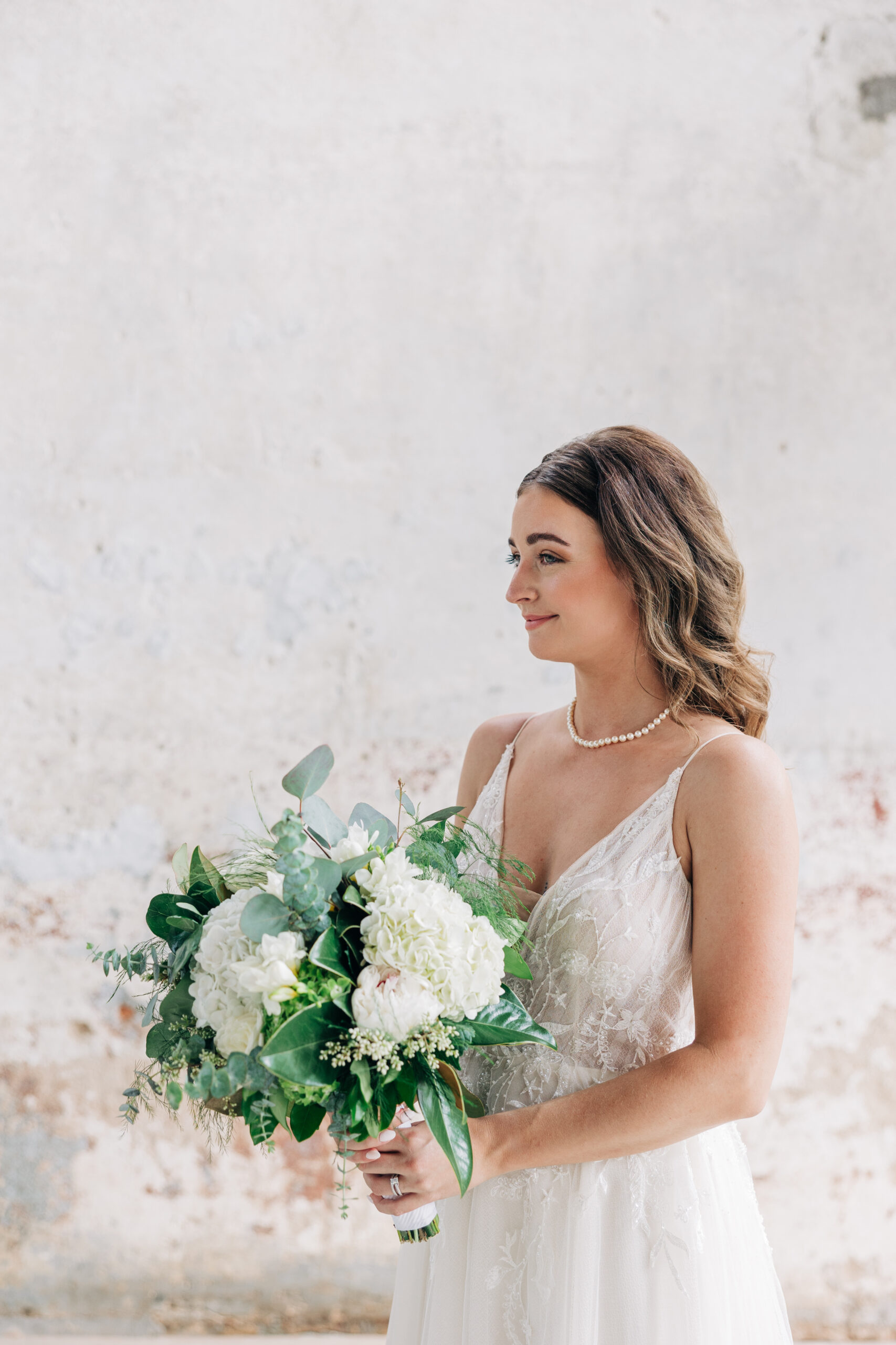A bride smiles while holding her white bouquet in a lace dress in a rustic building