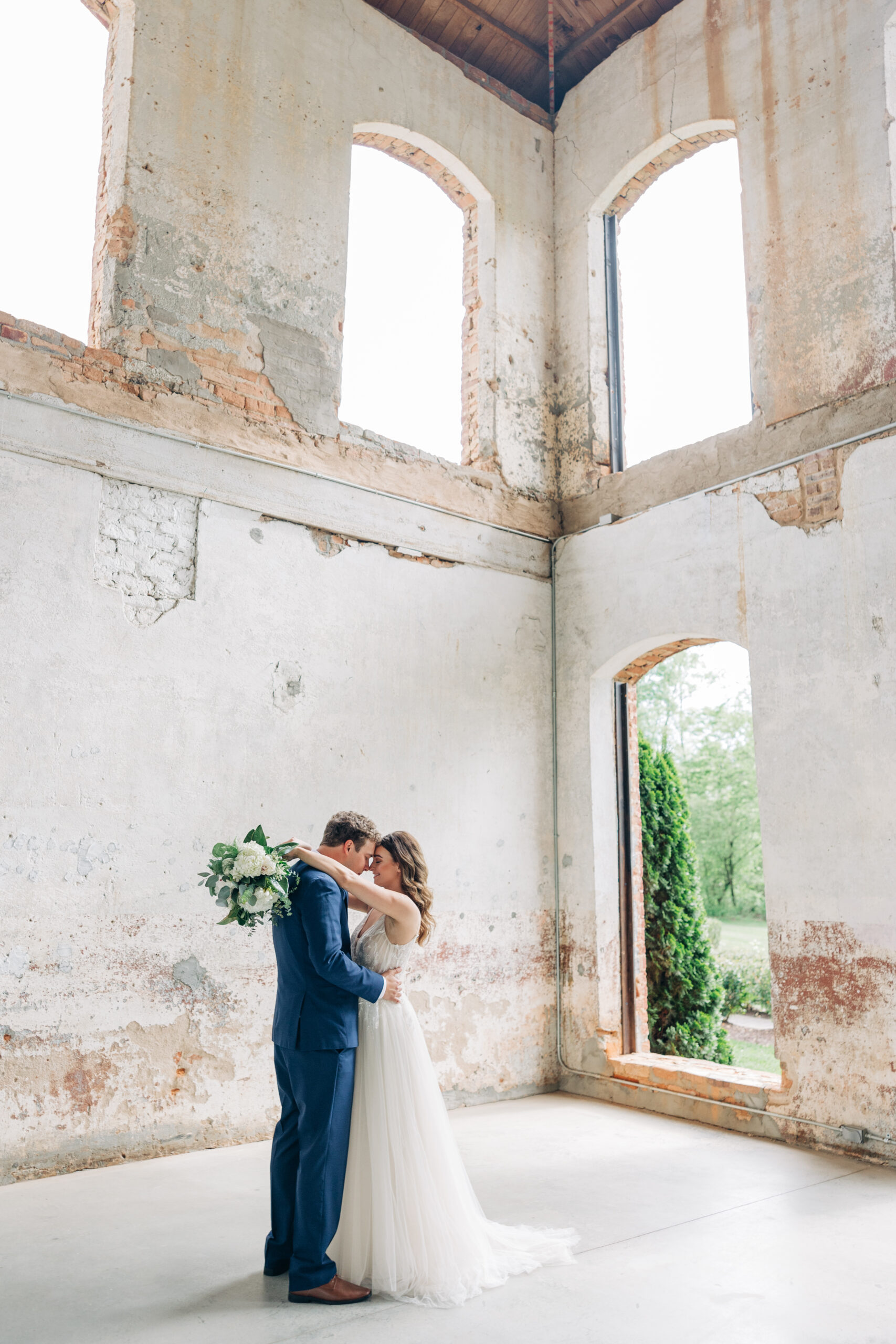 A groom in a blue suit dances closely with his bride in a rustic building
