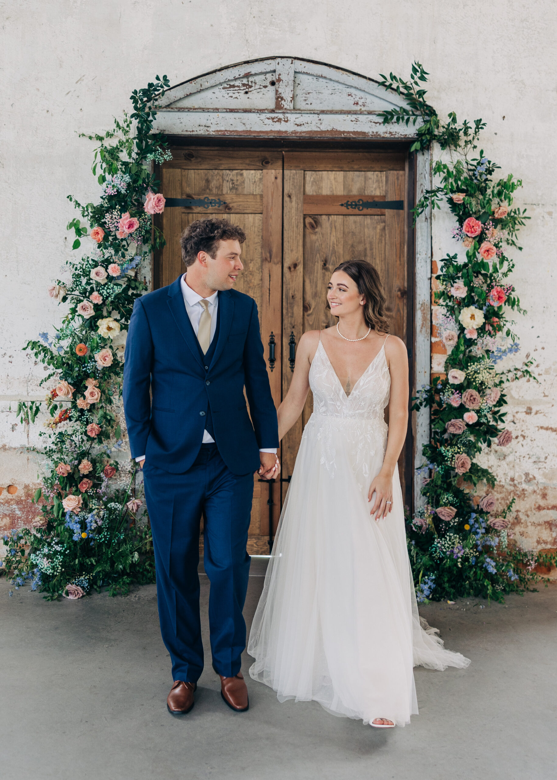 Newlyweds hold hands in front of a large wooden door covered in colorful flowers and walk