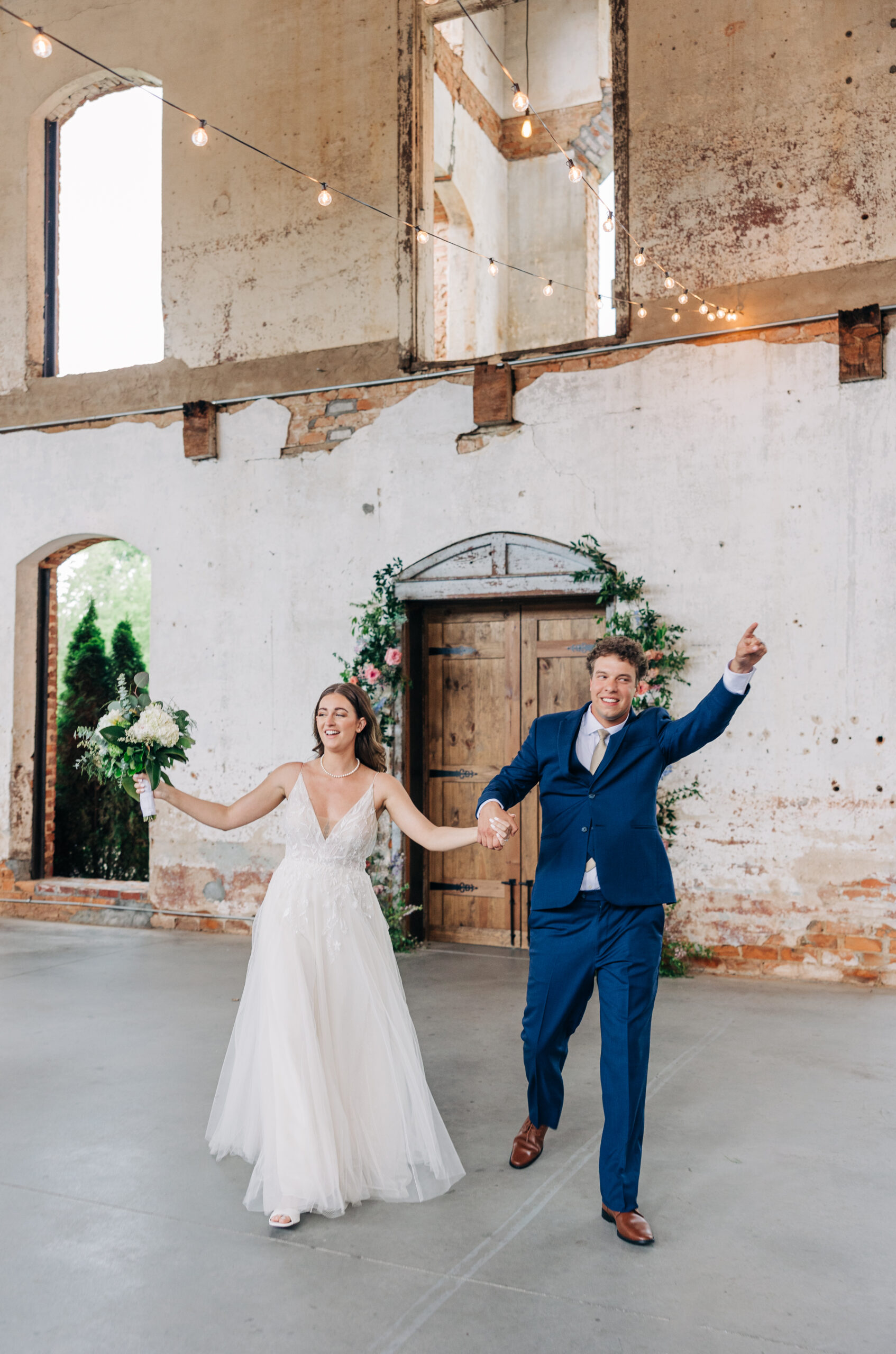 A bride and groom in a lace dress and blue suit enter their wedding reception with arms up and big smiles