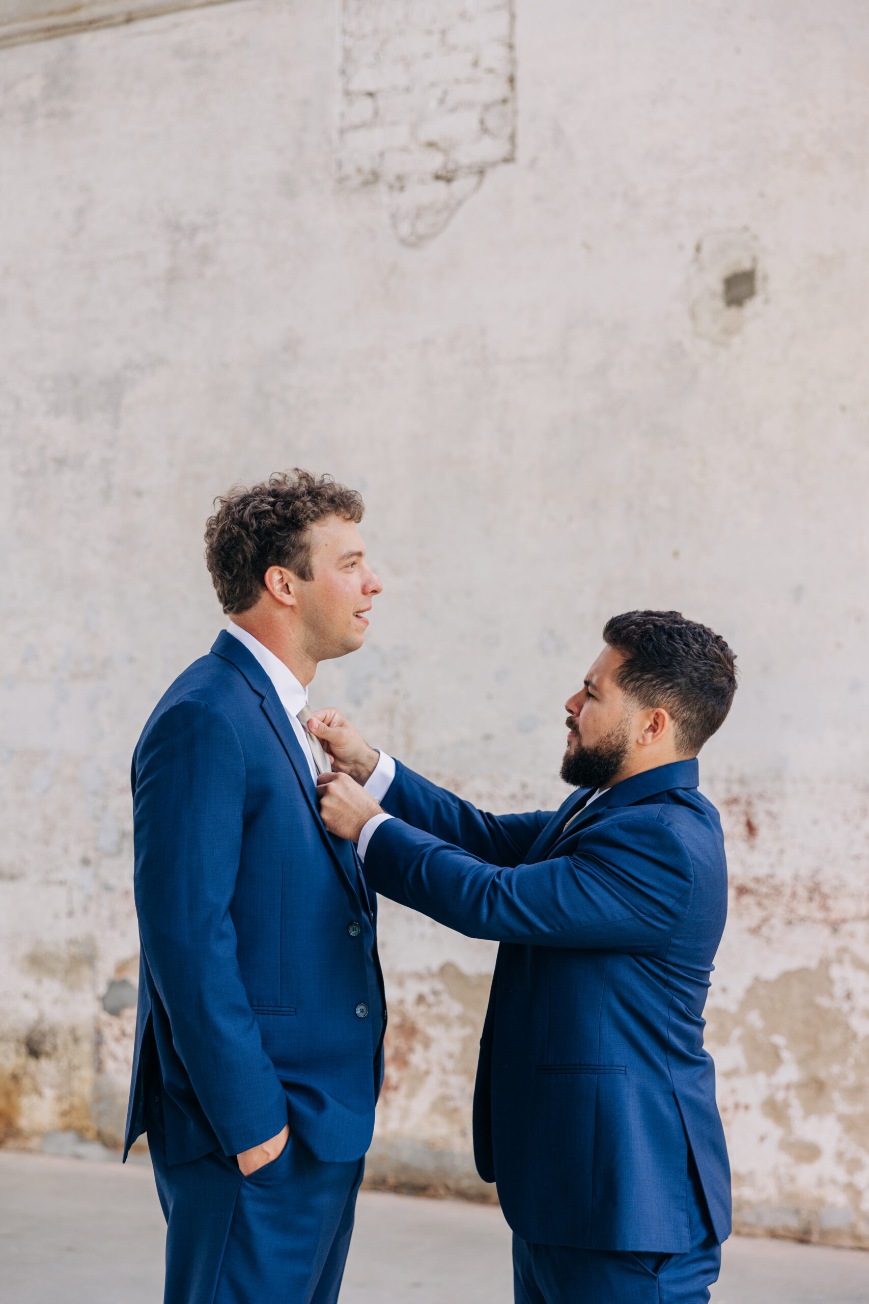 A groomsmen in a blue suit helps the groom straighten his tie in a rustic building