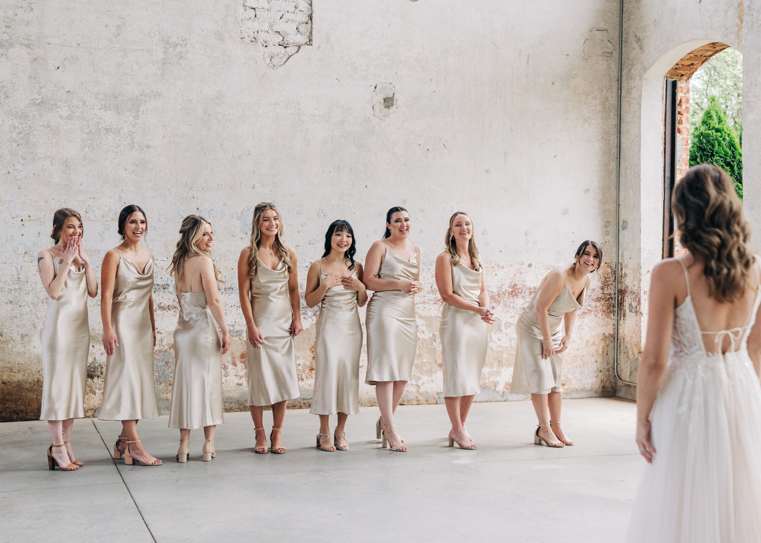A bride shows off her dress and look to her bridesmaids in a rustic building