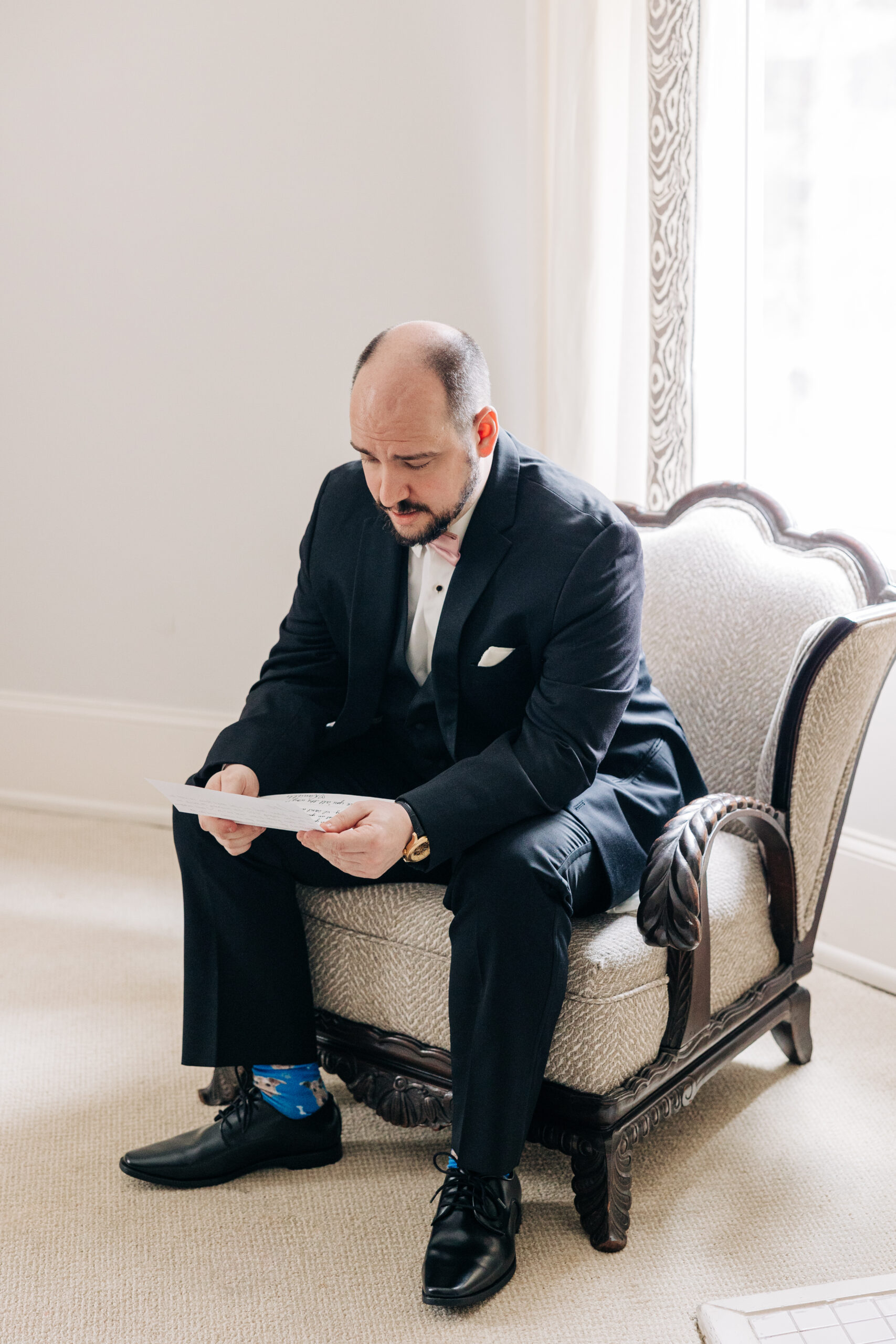 A groom sits in an ornate chair under a window reading a letter in his suit