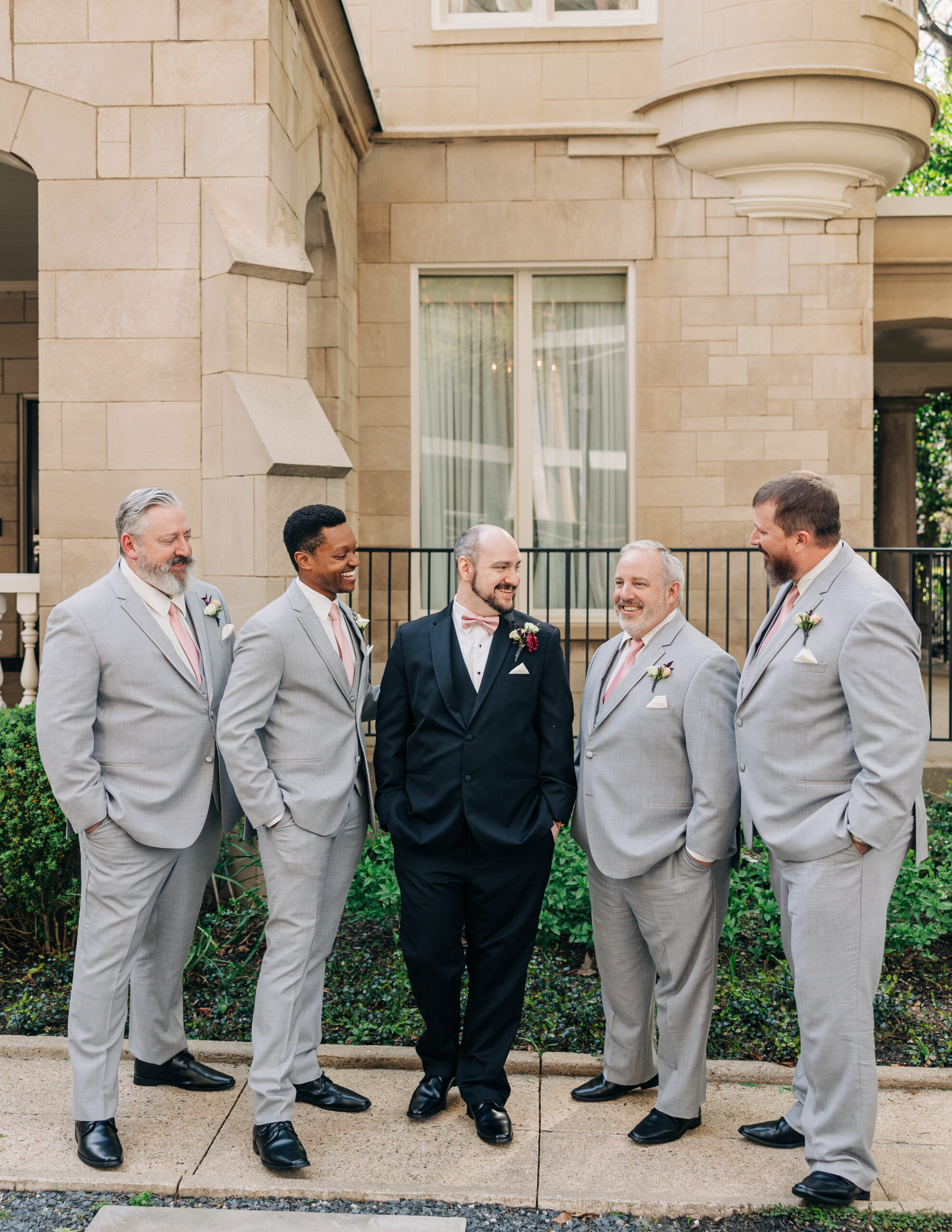 A groom stands on a sidewalk laughing with his groomsmen in grey suits
