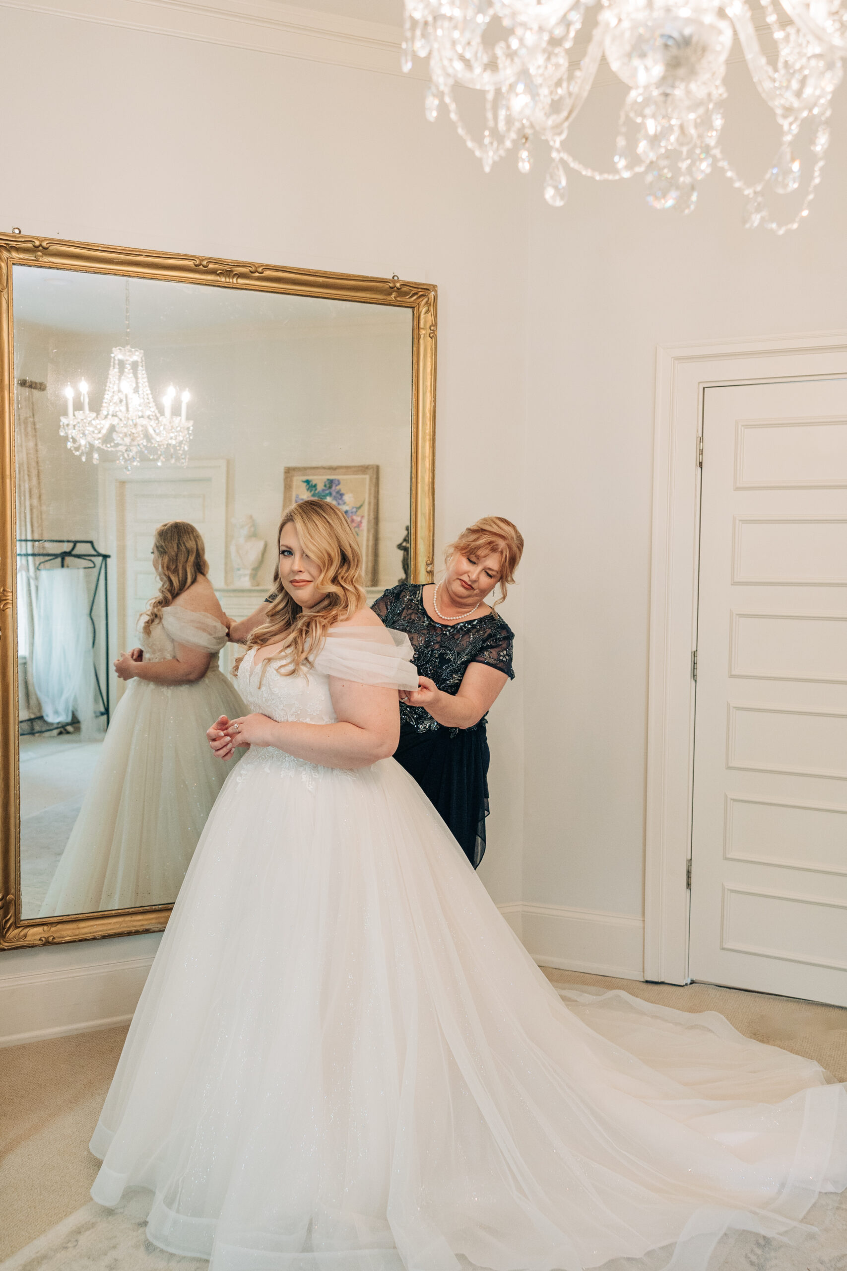 A bride is helped by mom to finish getting ready in a large mirror at her Wimbish House wedding