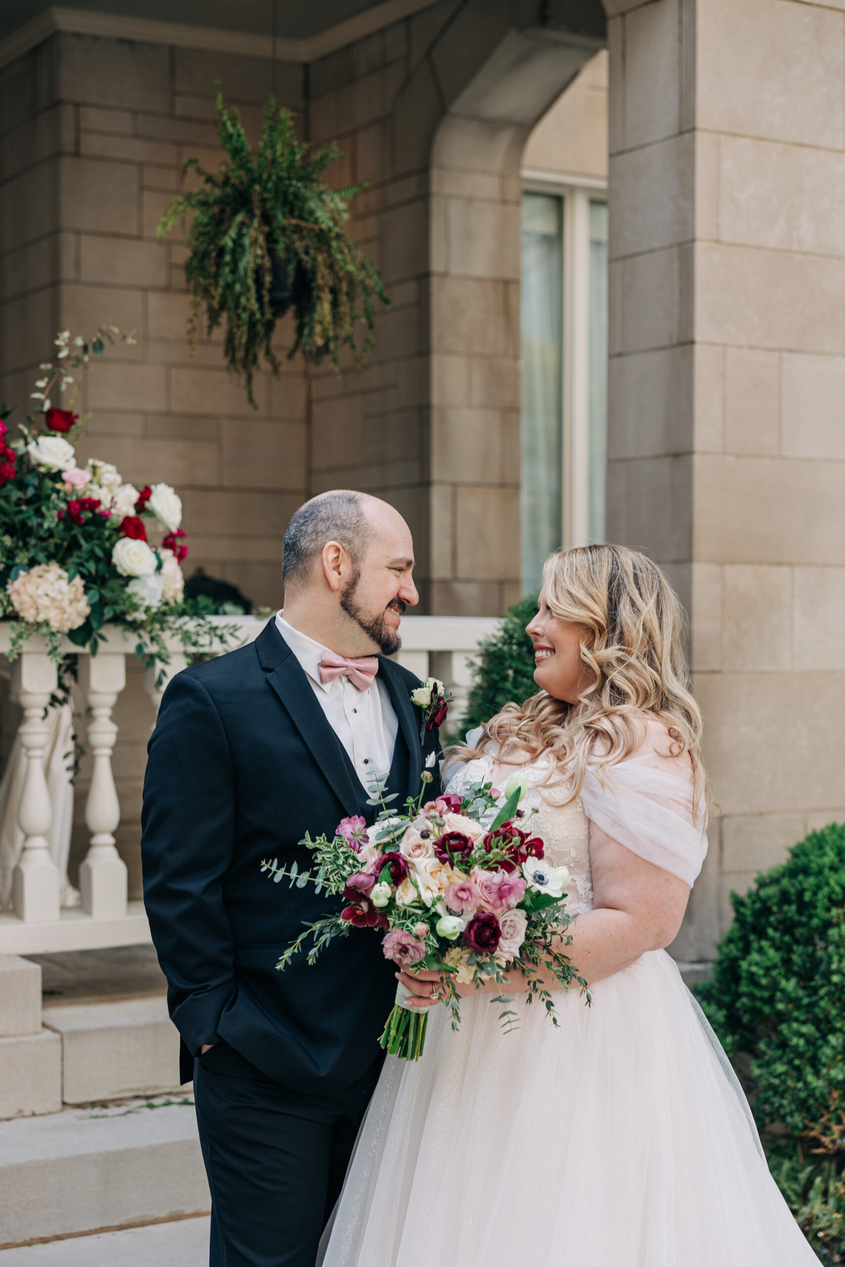 Newlyweds laugh with each other while standing outside the front porch holding the colorful bouquet