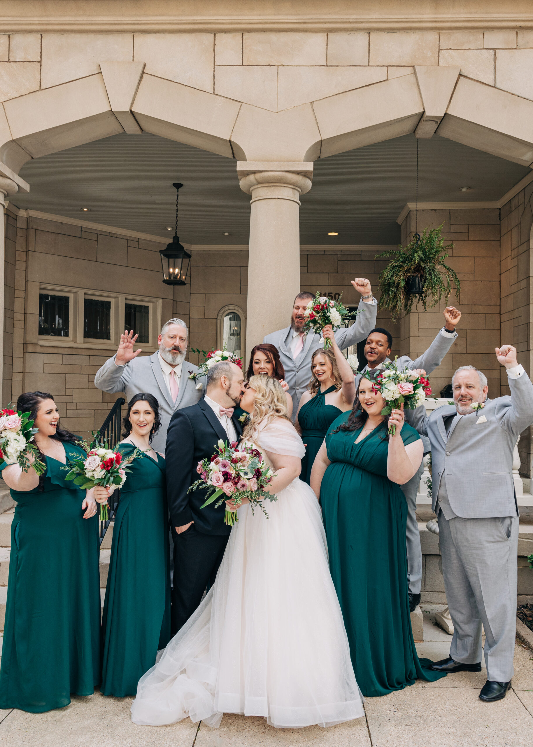 Newlyweds kiss on the front steps of their venue with their wedding party celebrating