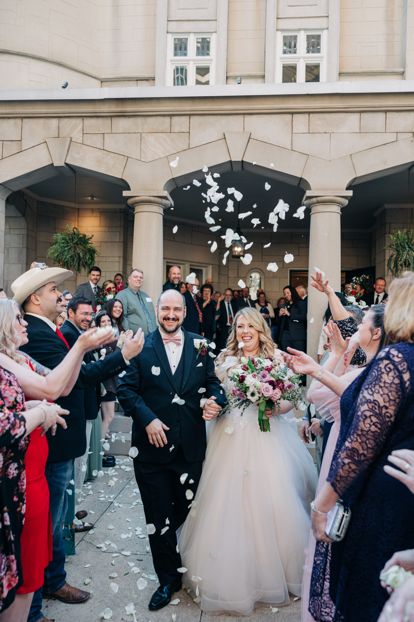 Newlyweds exit their wedding under a shower of white rose petals thrown by their guests