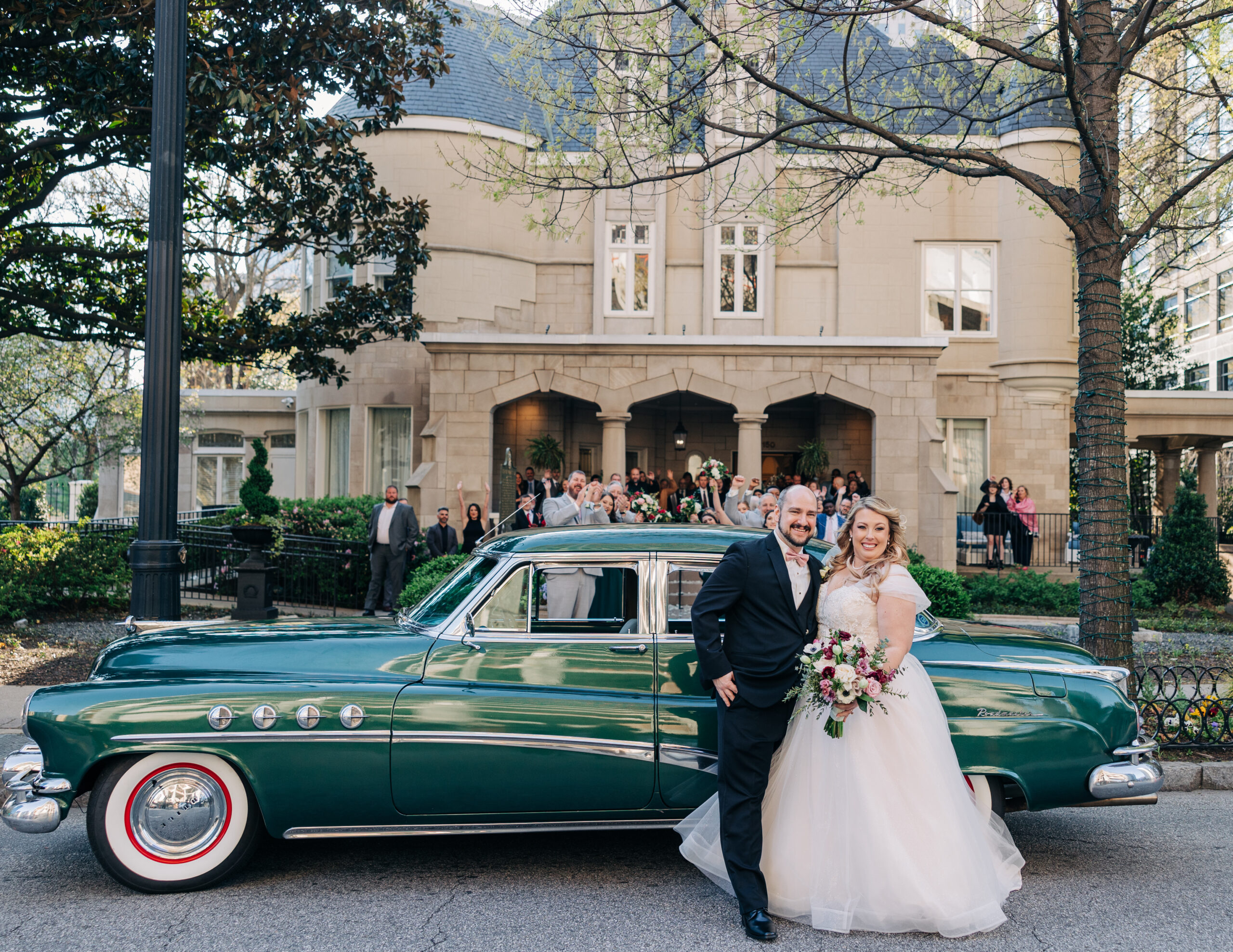 Newlyweds stand in front of a green vintage car with their Wimbish House wedding guests cheering